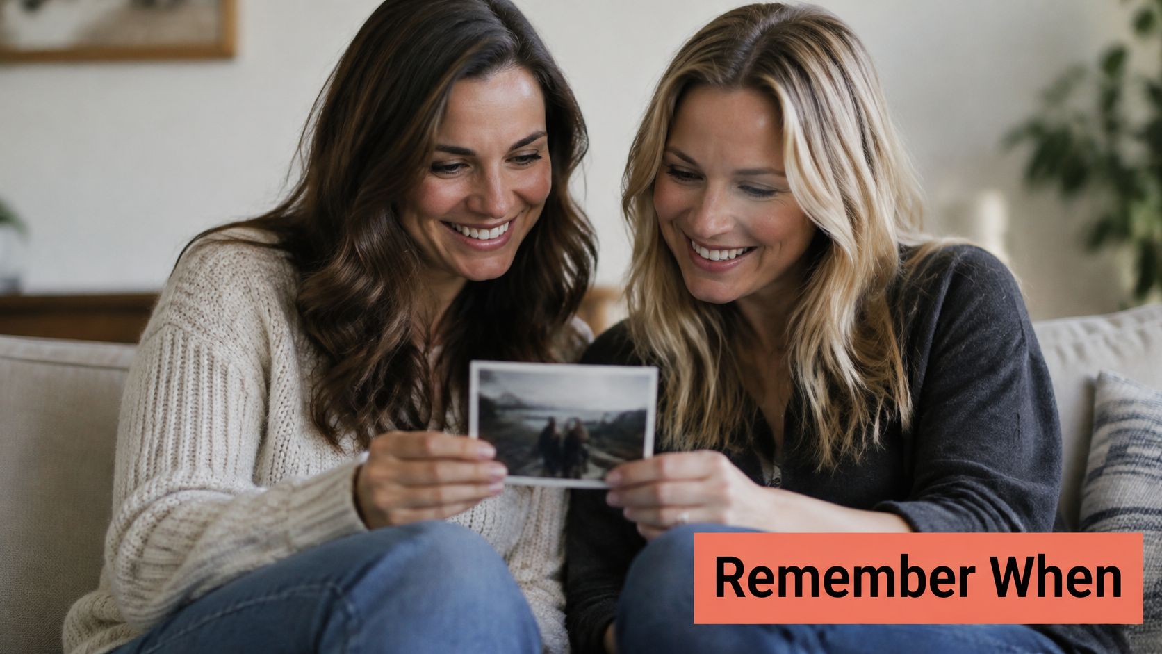 Two happy female friends sitting on a couch together while looking at a nostalgic printed photograph.