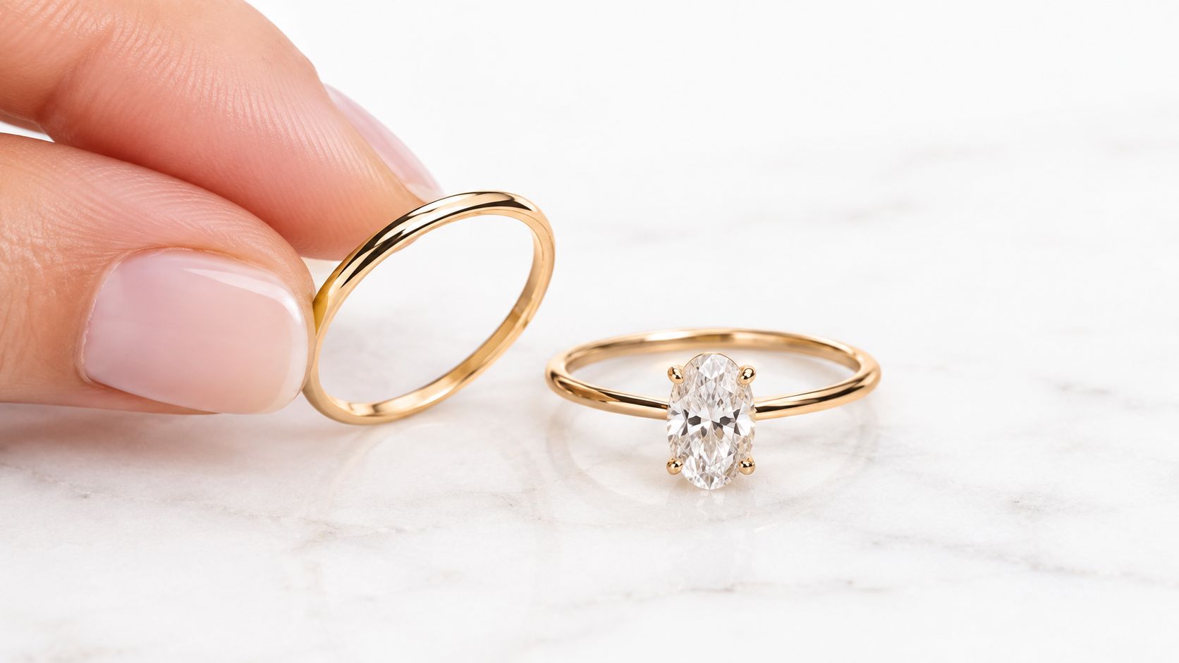 A hand holding a thin gold wedding band next to a gold oval diamond engagement ring.