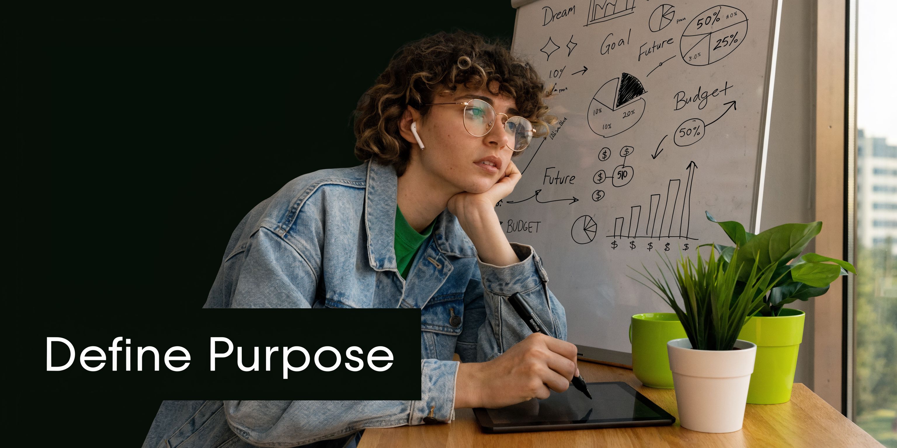 A focused young woman in a denim jacket sketching on a tablet in a modern office workspace.
