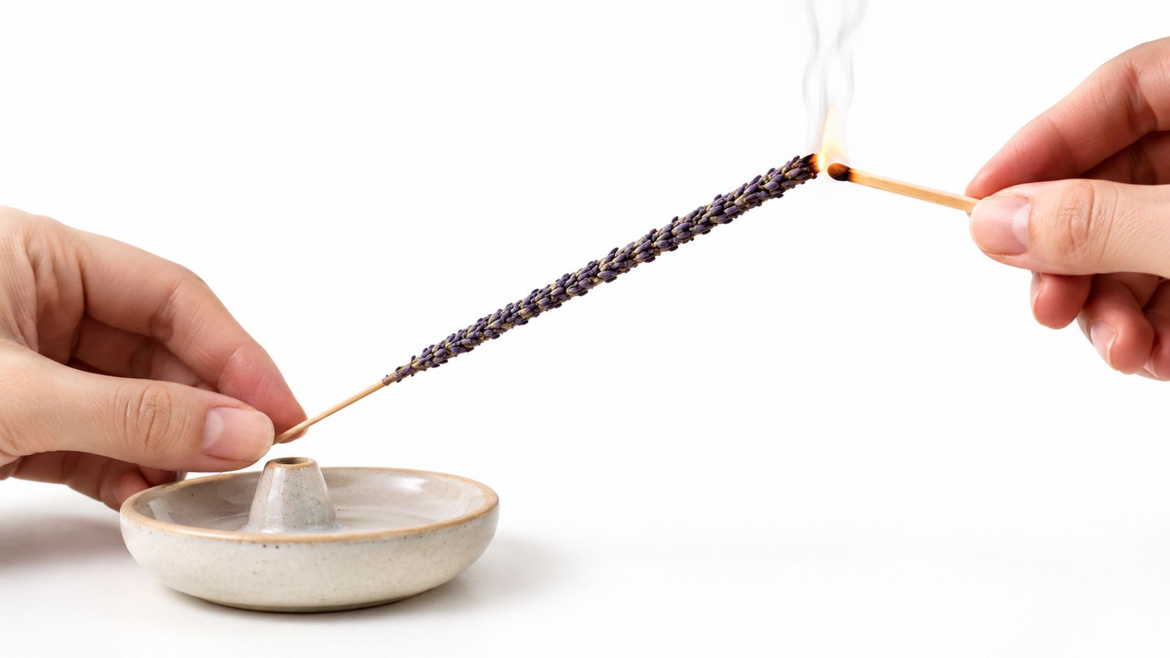 A hand holding a match to light a dried lavender incense stick held over a ceramic tray.