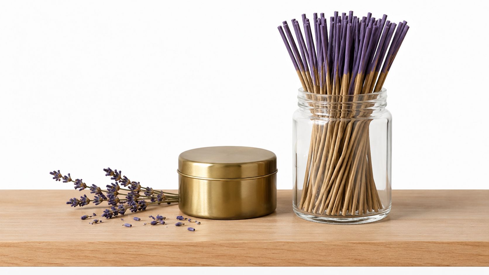 A glass jar filled with lavender incense sticks stands next to a gold metal container and lavender sprigs.