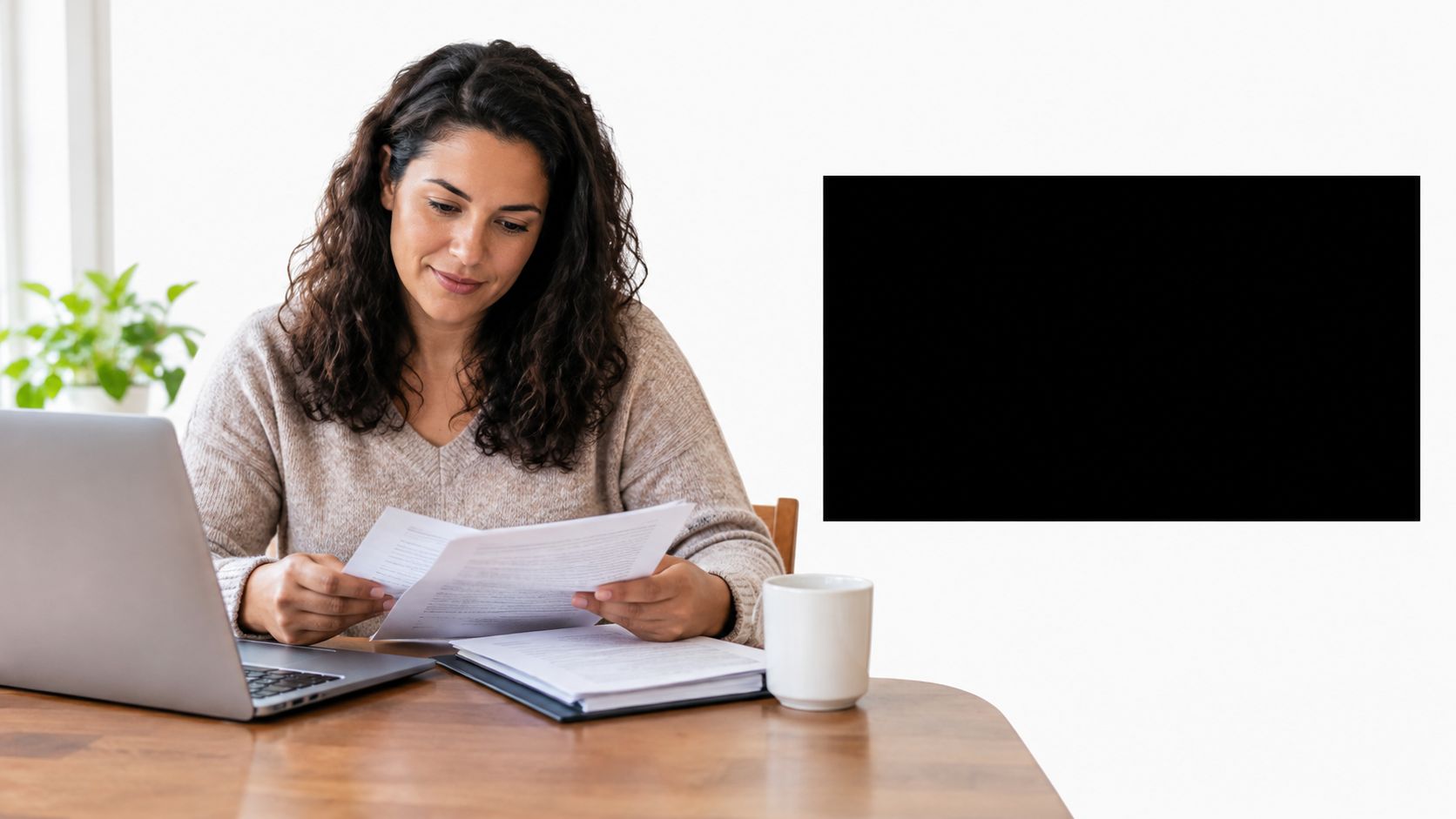 A woman reviewing loan documents while working on her laptop at a desk at home.