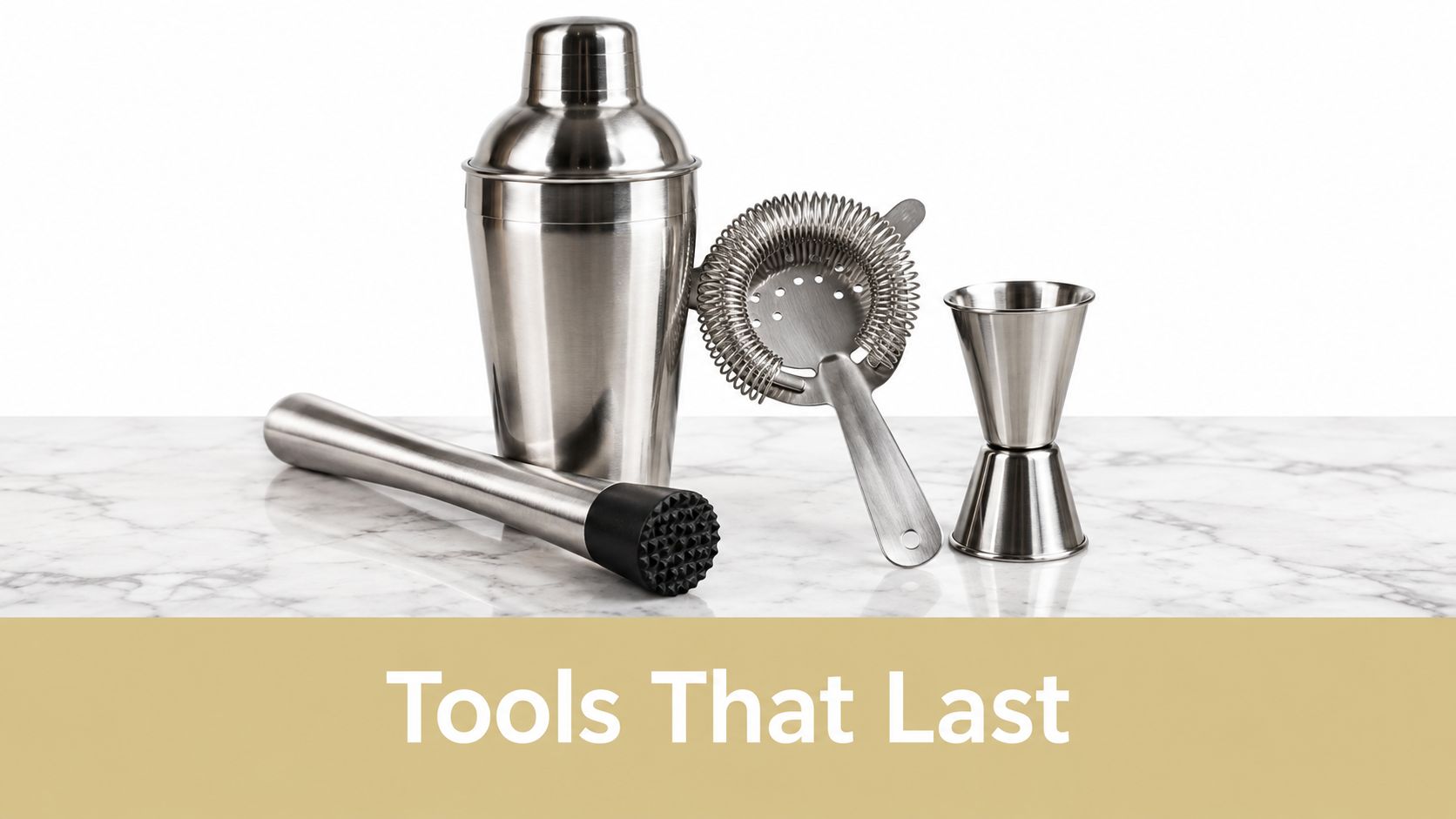 A stainless steel cocktail shaker set including a muddler, strainer, and jigger on a white marble surface.