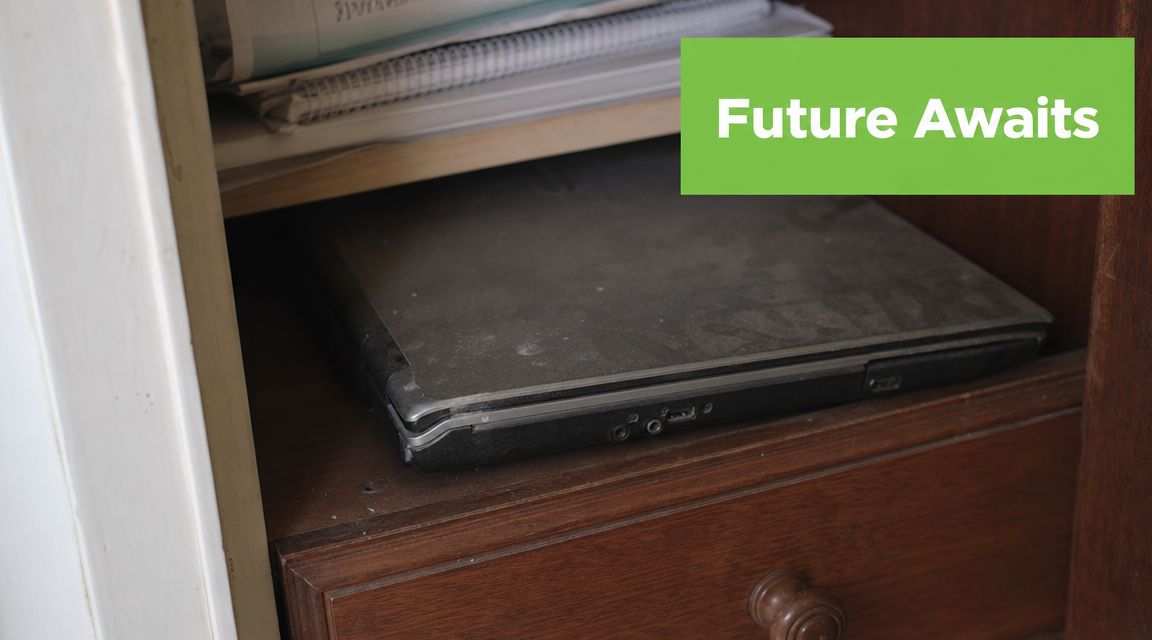 A dusty, closed laptop sits on a wooden shelf beneath some papers in an old desk.
