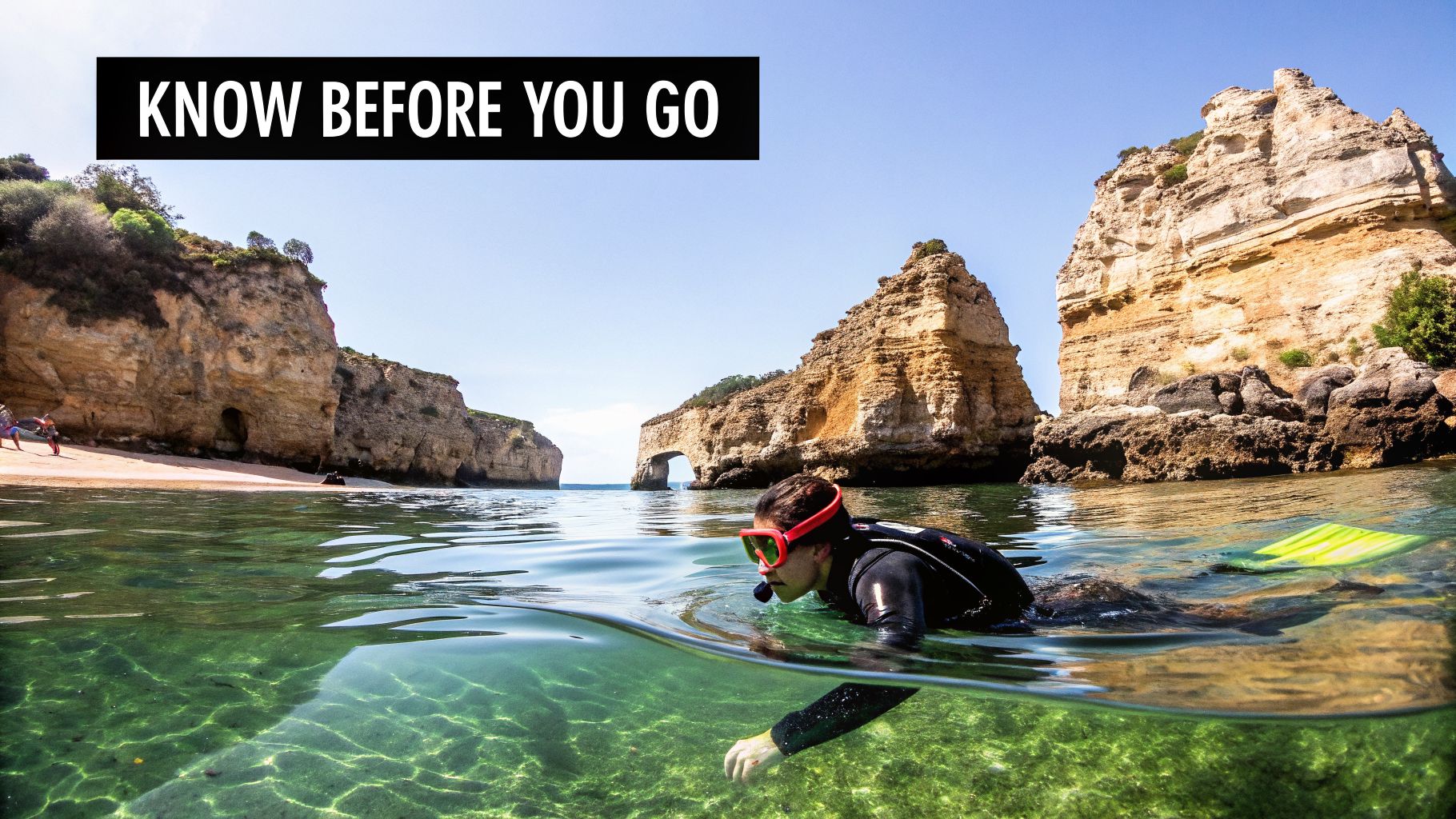 A person snorkeling in clear green water with dramatic cliffs and a natural arch in the background.