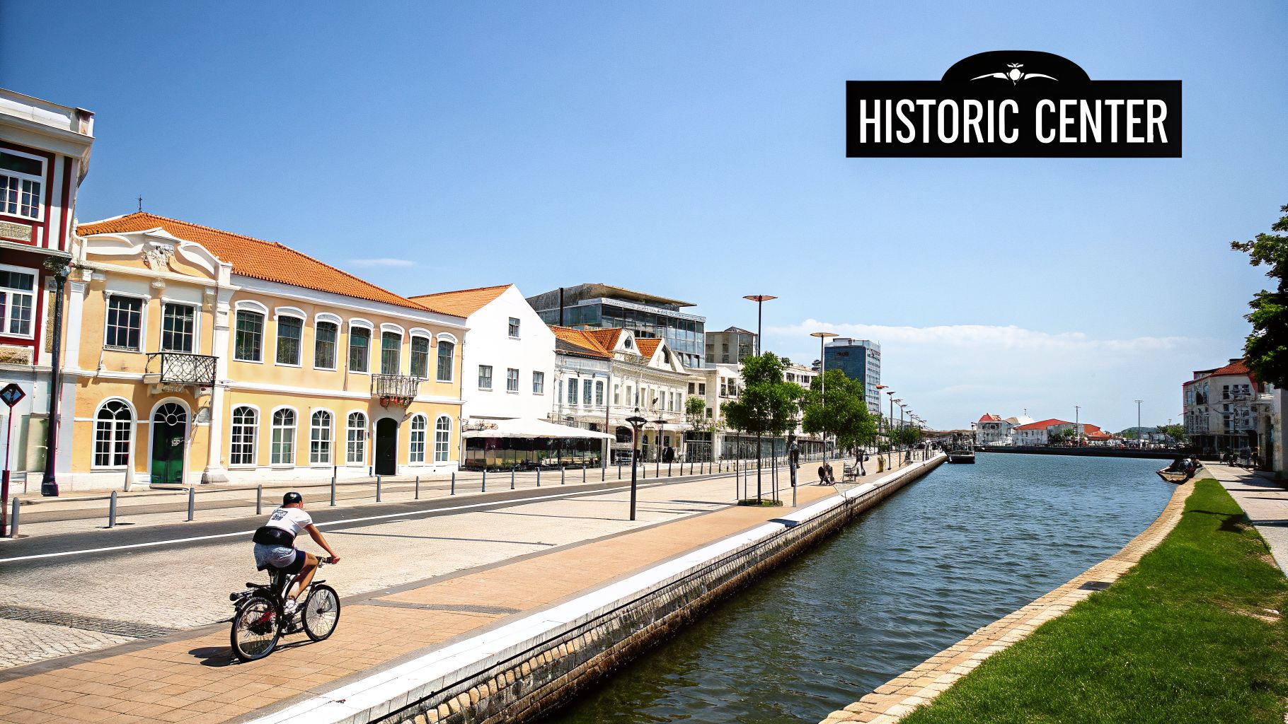 A cyclist rides past colourful historic buildings along a scenic canal in Aveiro, Portugal under a clear blue sky.
