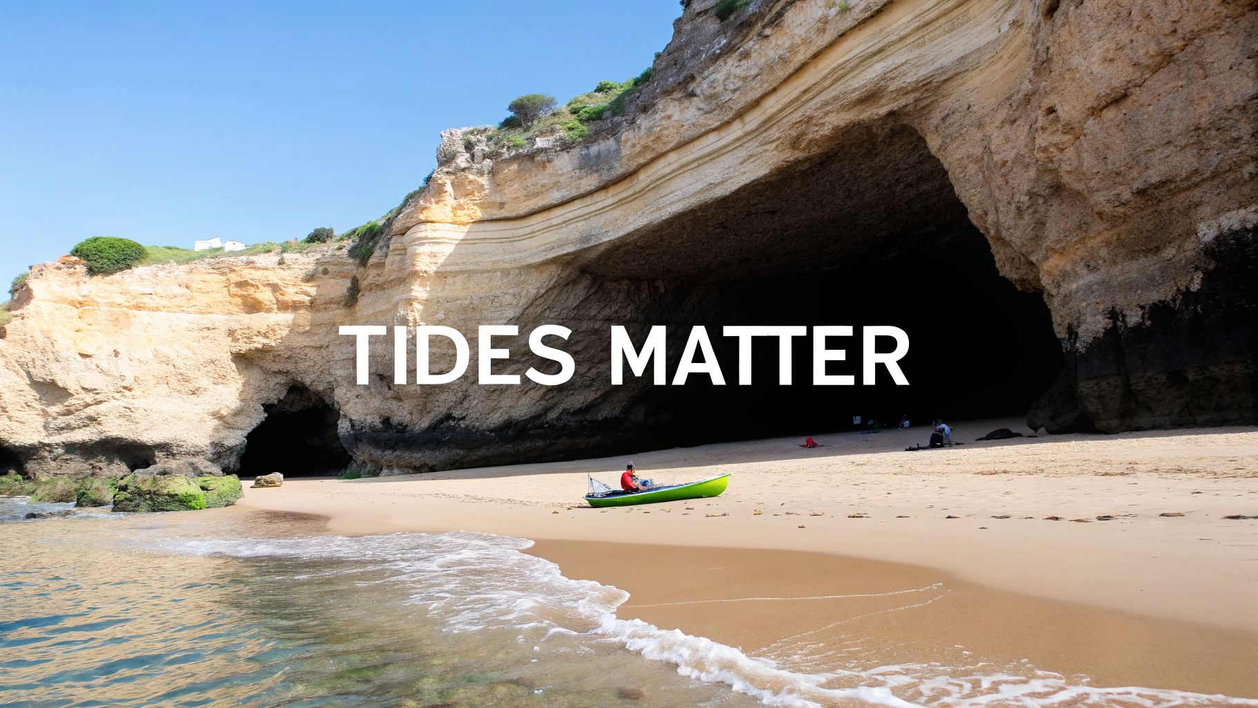 A sunny beach with a large sea cave entrance and a person sitting in a green kayak on the sand.