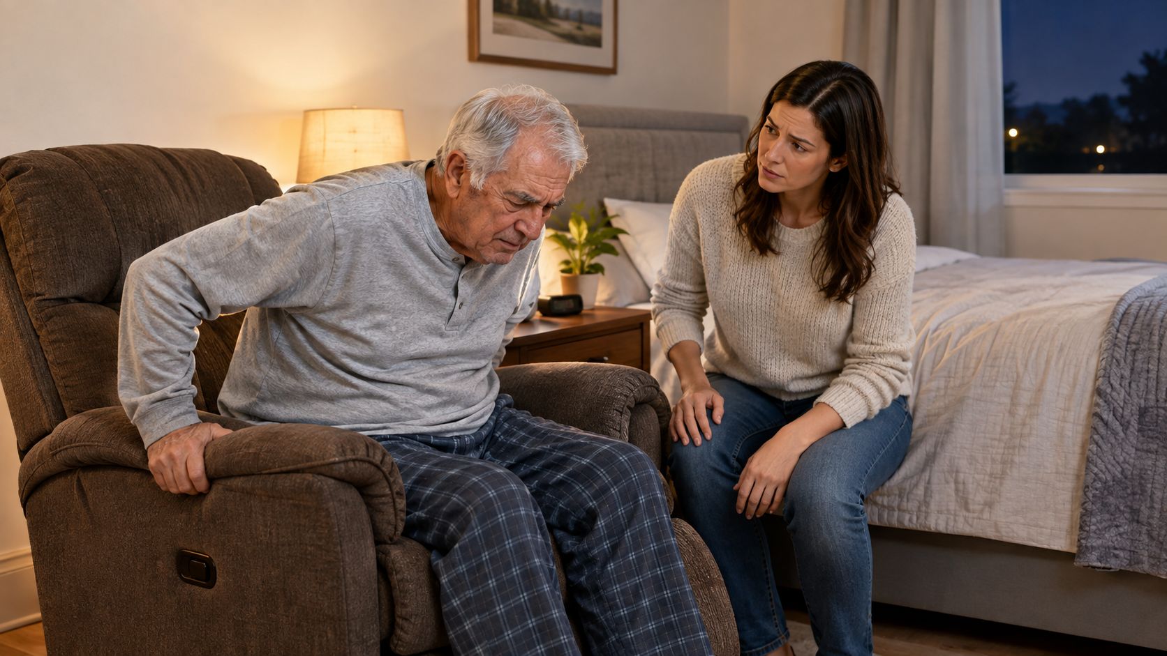 A concerned woman comforts an elderly man struggling to stand up from a recliner chair in his bedroom.