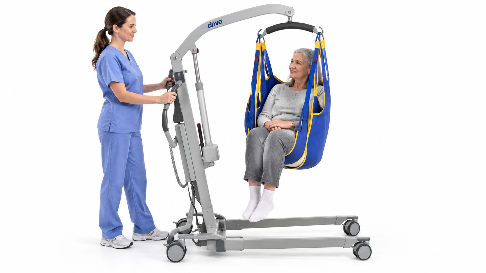 A caregiver in blue scrubs operates a patient lift device to support a woman seated in a sling.