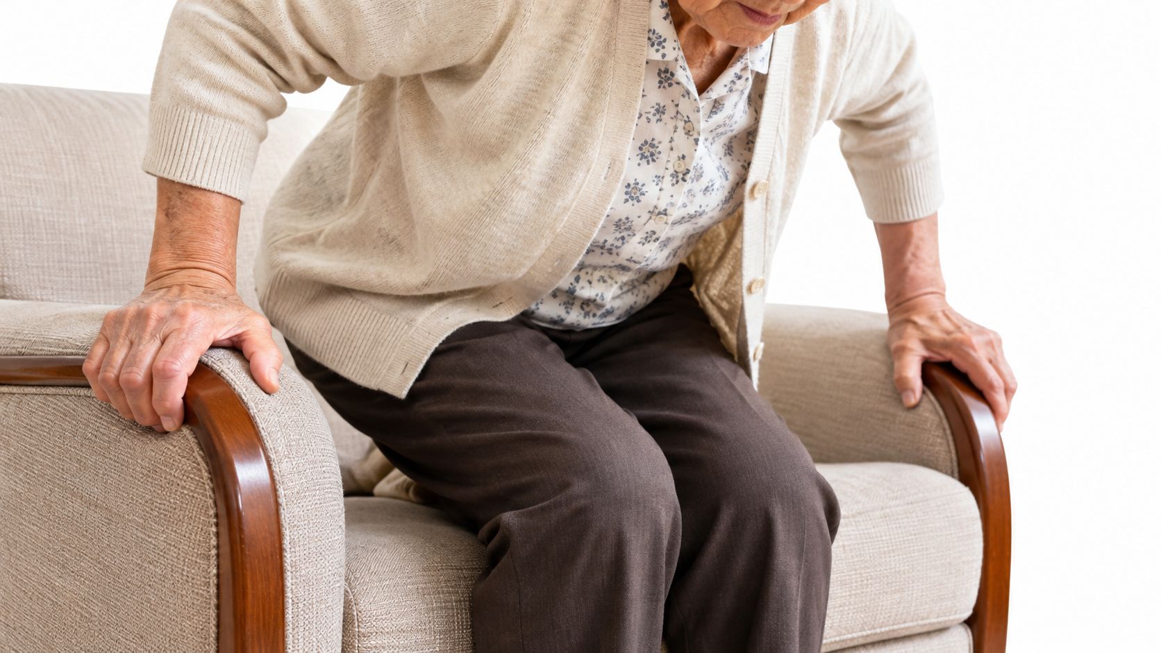 An elderly person struggling to stand up from a comfortable beige armchair by pushing on armrests.