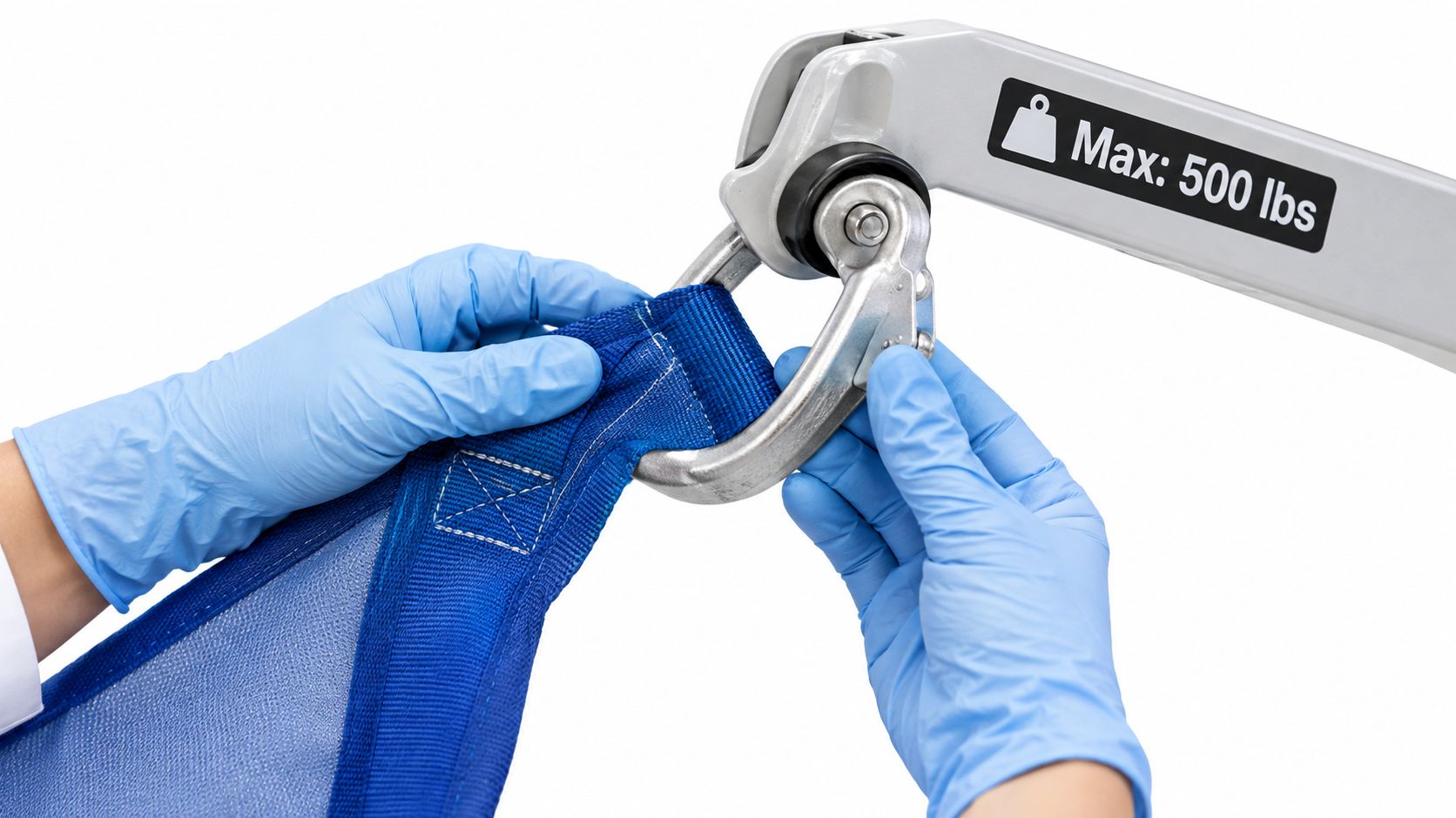 A caregiver wearing blue medical gloves attaches a blue patient sling strap to a medical hoist hook.