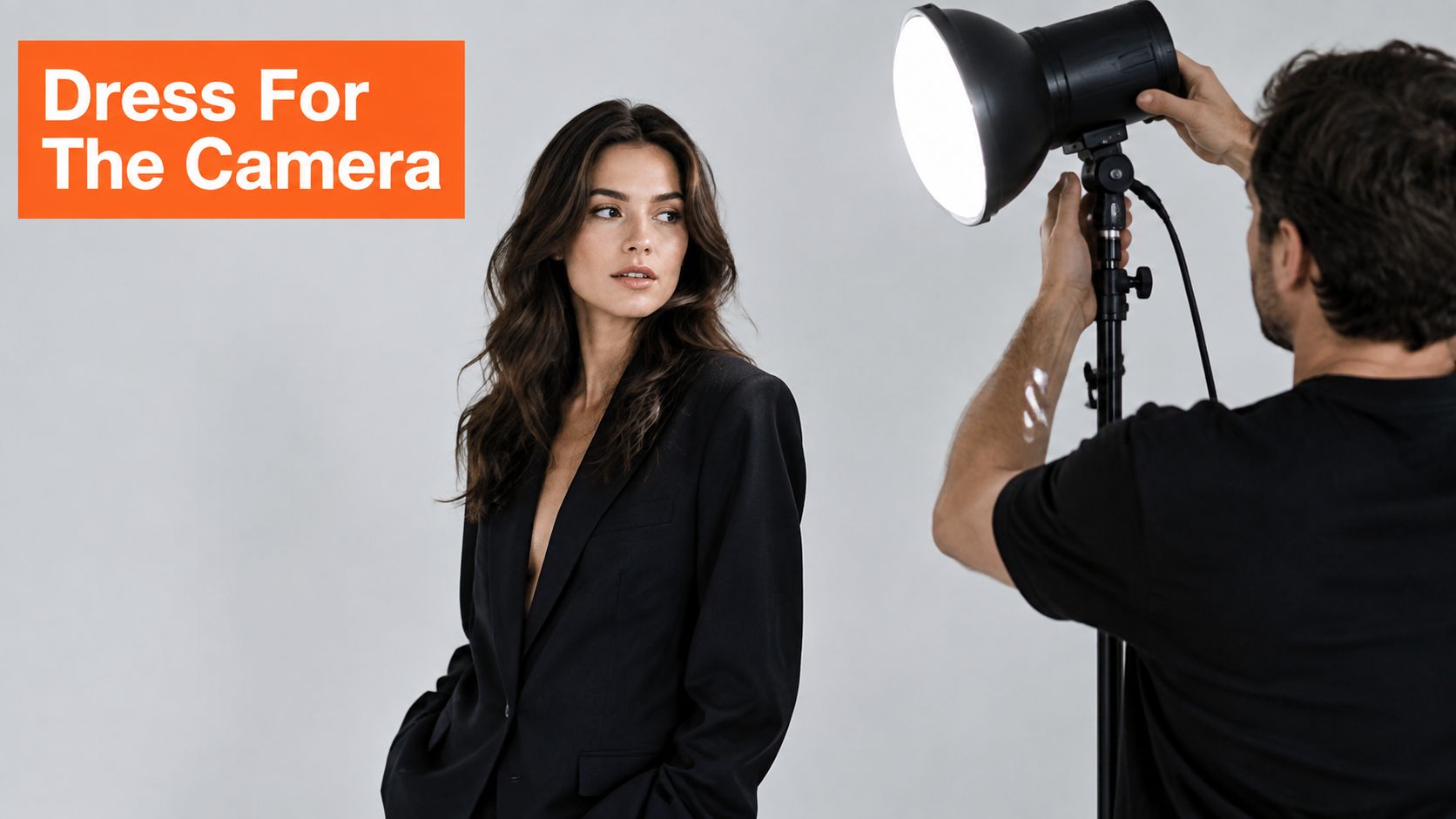 A professional photographer adjusts a lighting setup for a woman wearing a black suit during a portrait session.