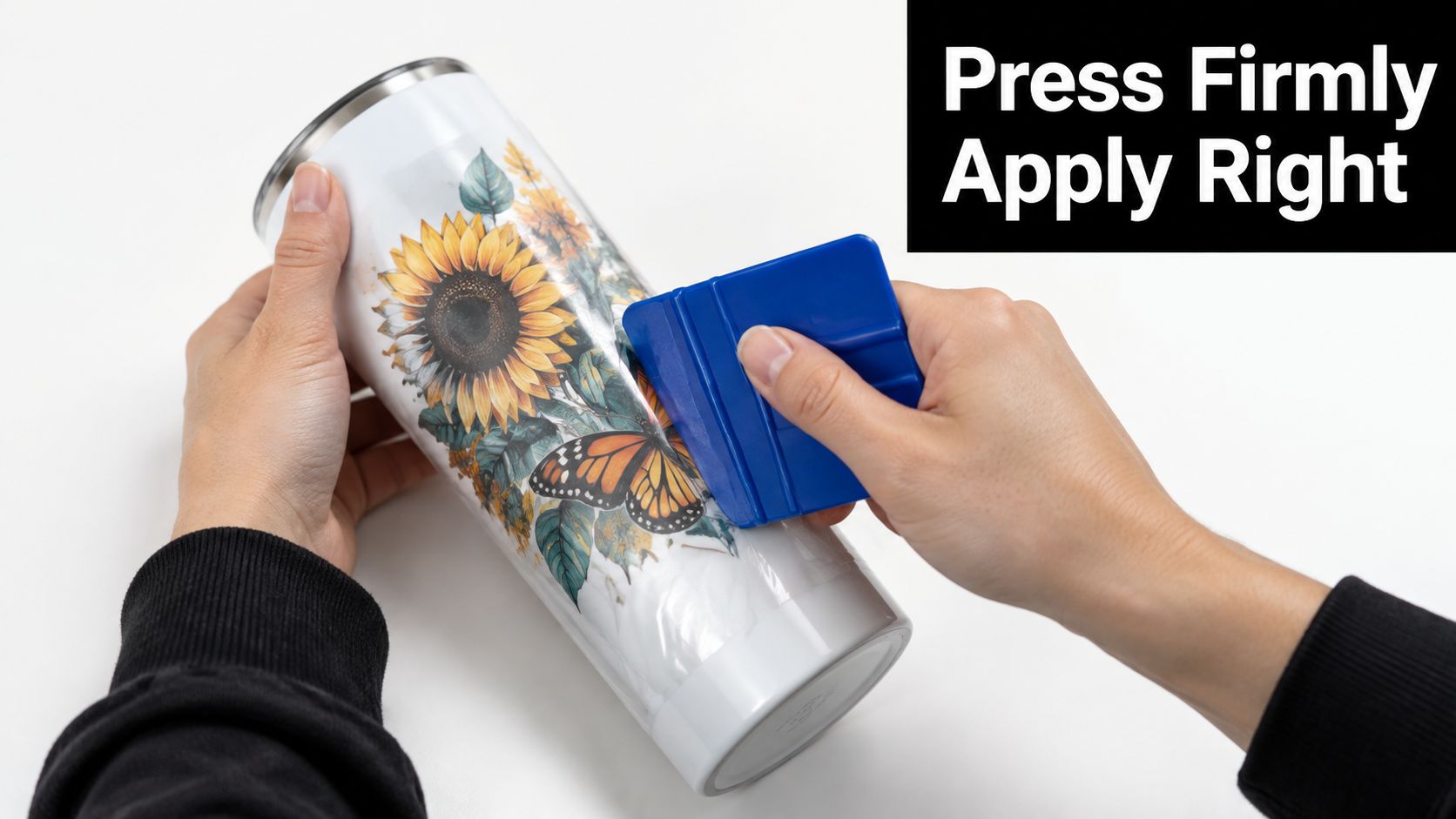 A person using a blue plastic squeegee to apply a decorative transfer onto a white tumbler.