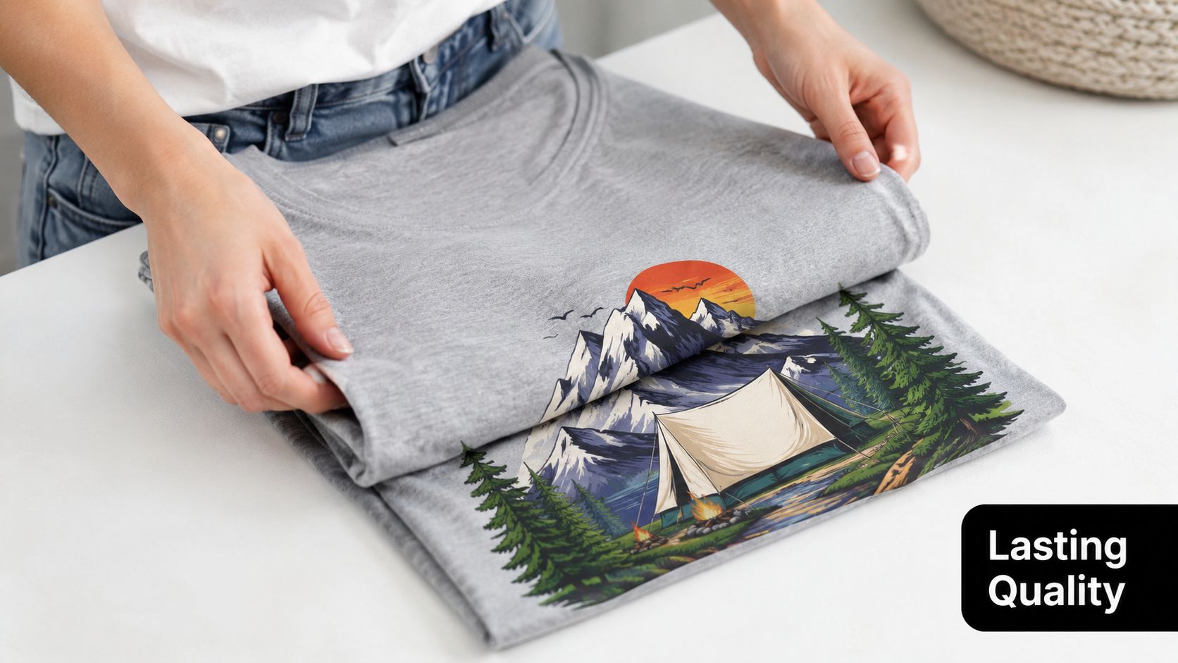 A person folding a grey t-shirt featuring a scenic camping design with mountains and a tent.