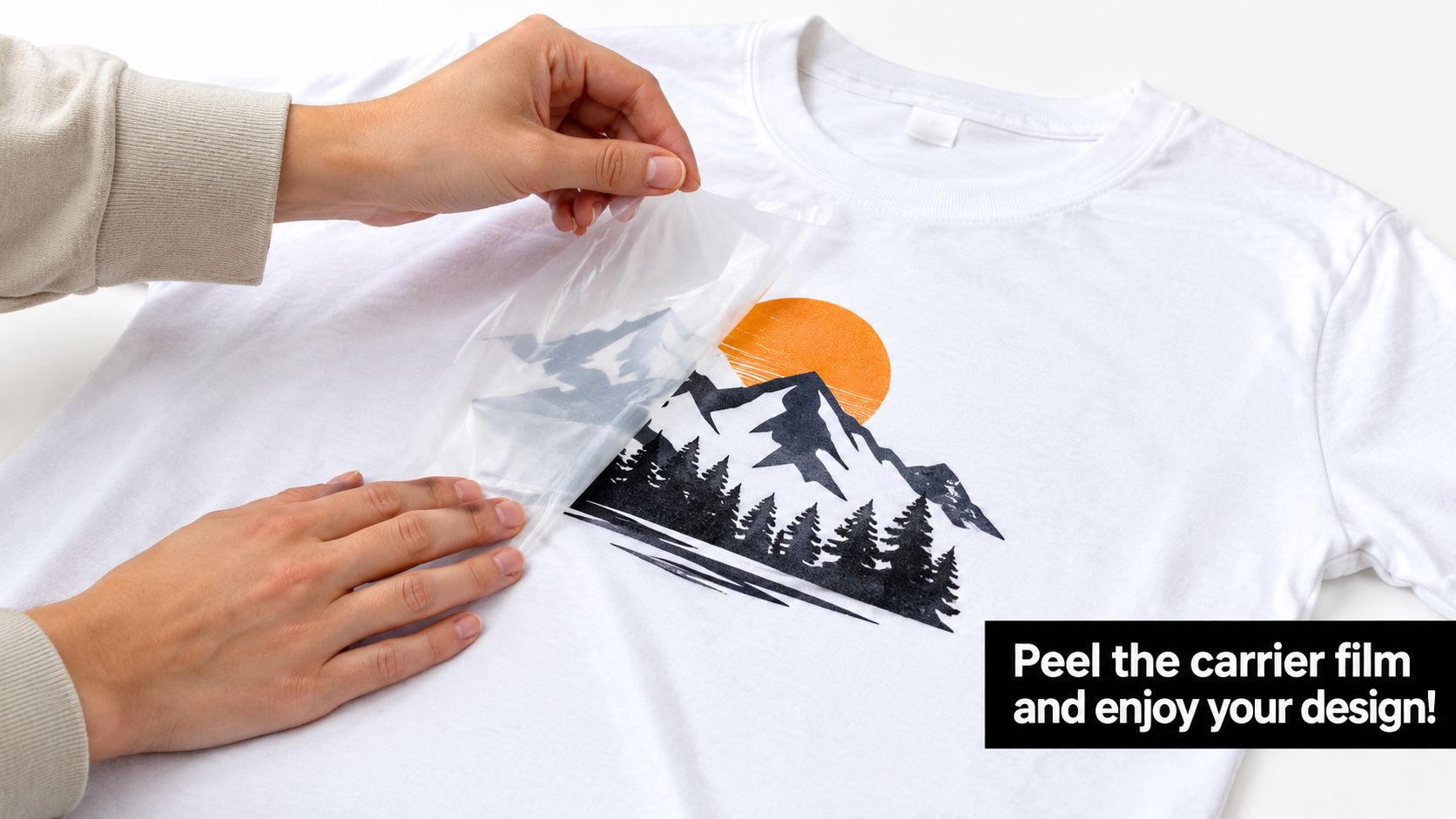 A person peels a transparent carrier film off a custom printed mountain design on a white t-shirt.