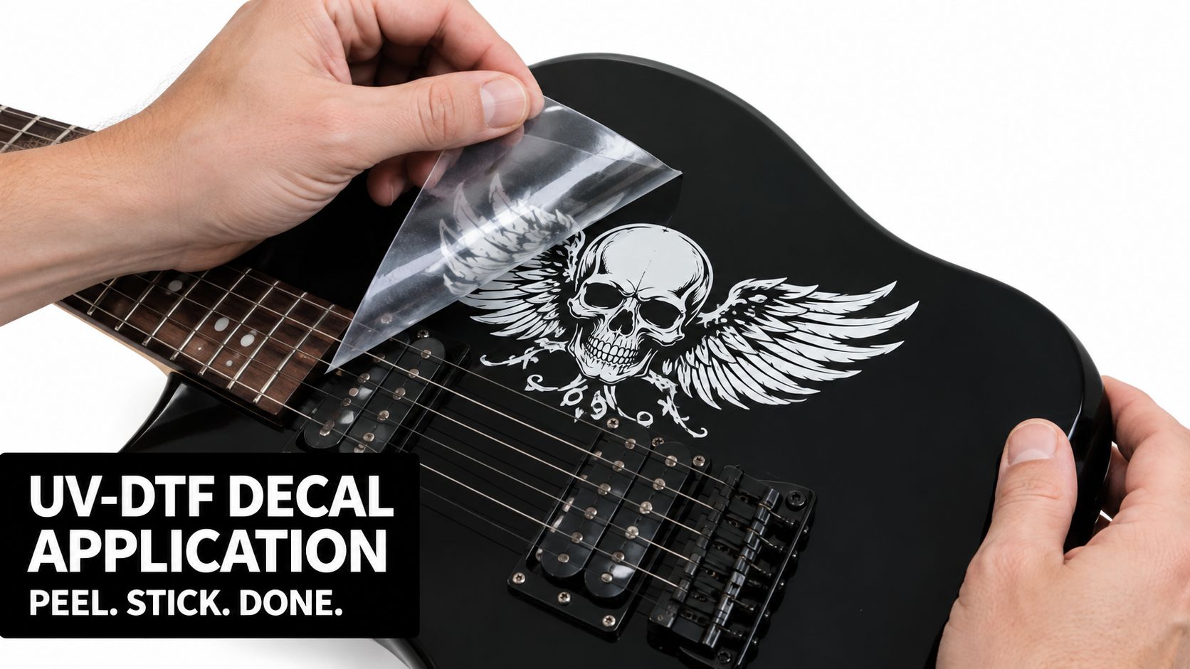 A close-up of a person peeling the clear backing off a skull and wings decal onto a guitar.