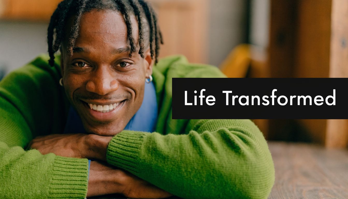 A smiling man with locs wears a green sweater and leans on his crossed arms, looking content.