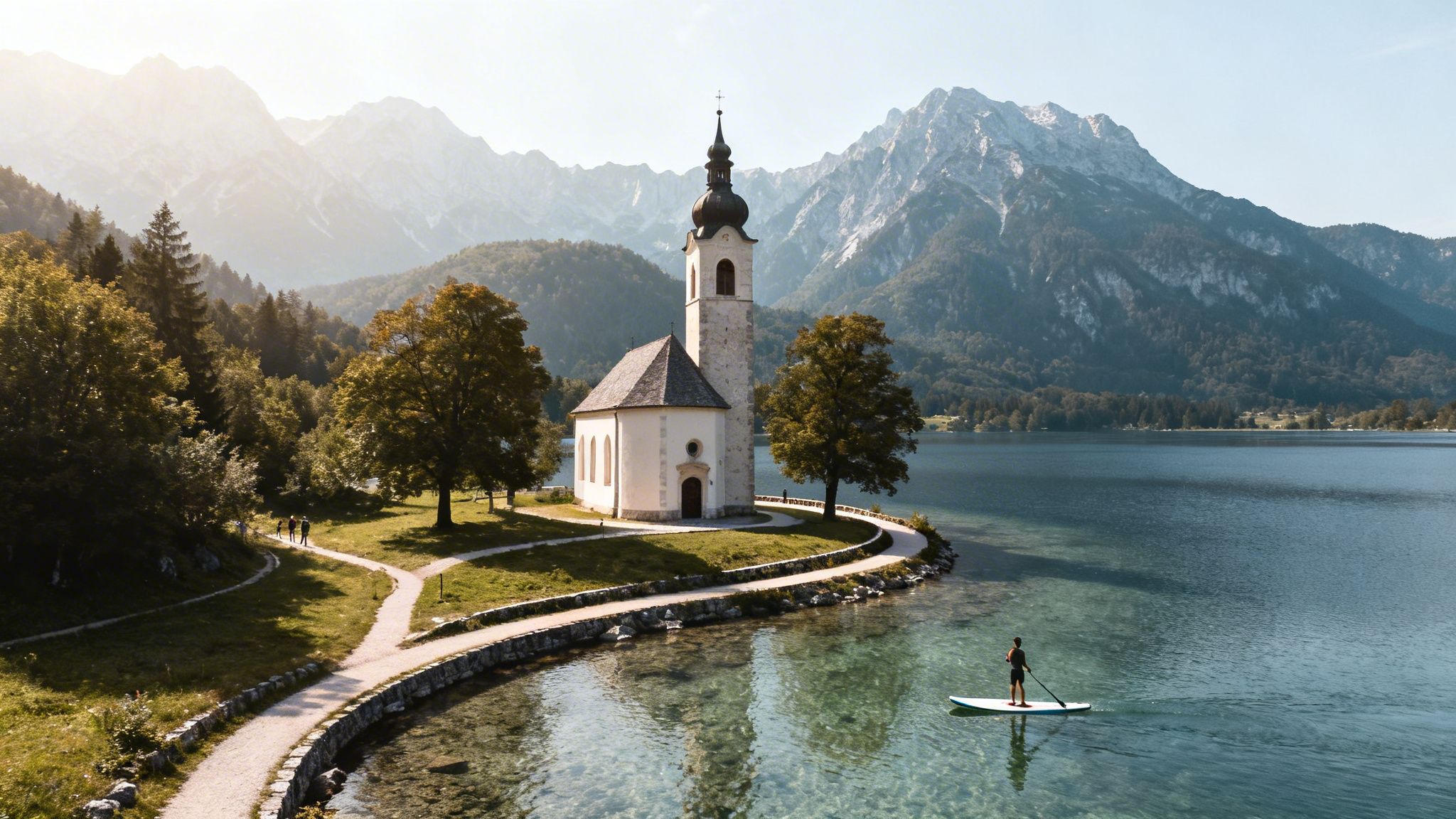A serene white church stands by a clear mountain lake, with a paddleboarder and hiking trails.