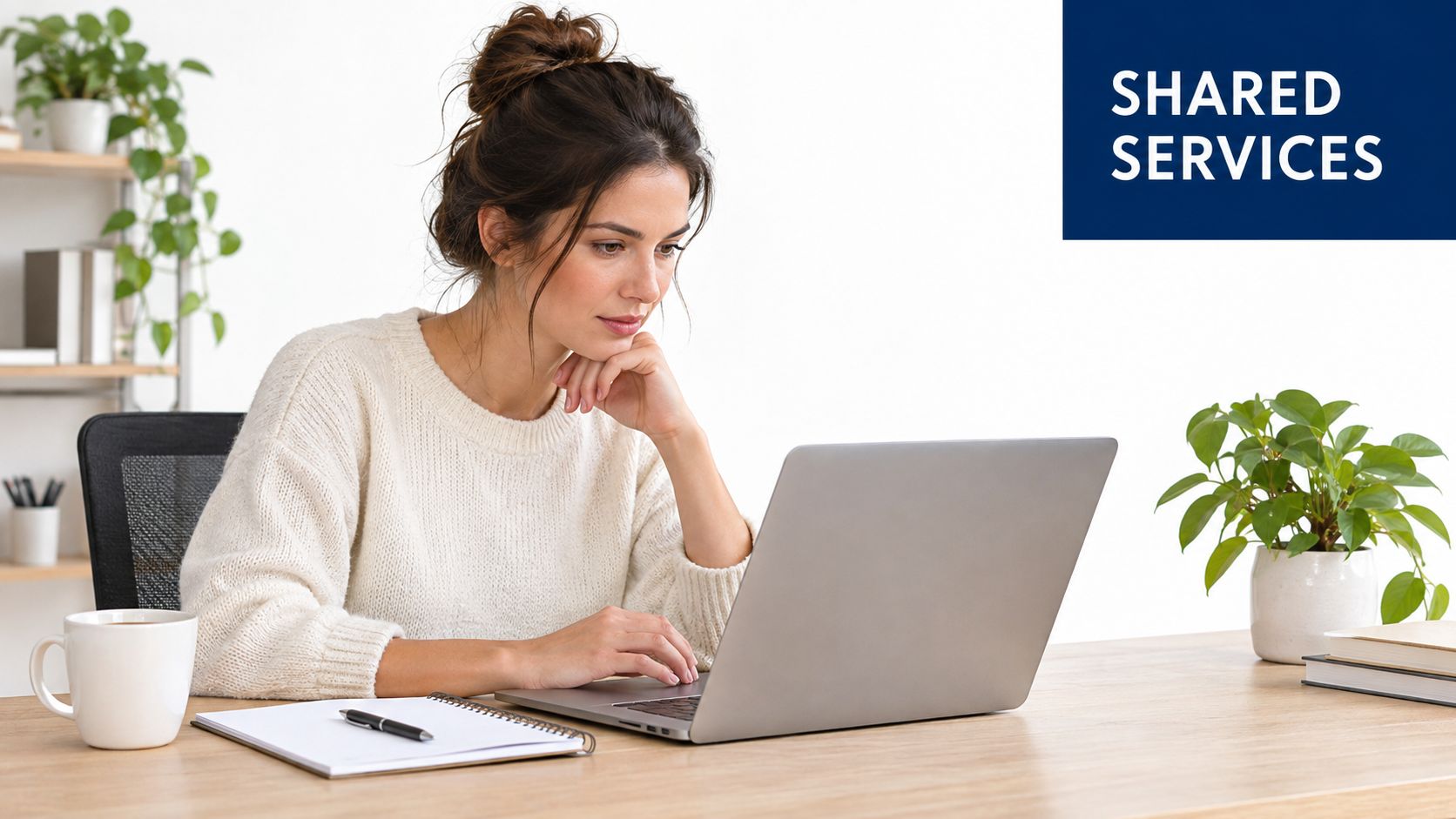 A woman working on a laptop at her desk with a notebook and coffee nearby.