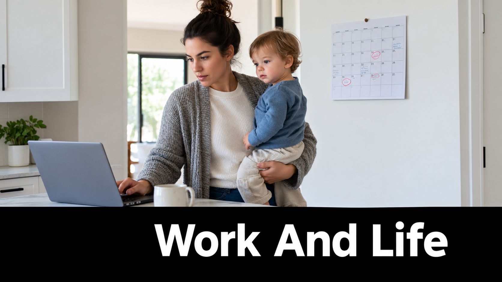 A mother works on her laptop while holding her young child in a bright home kitchen