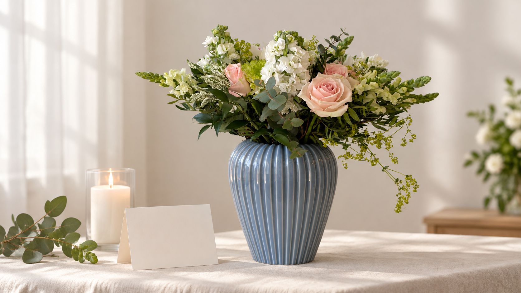 En smuk buket blomster i en blå vase står på et bord ved siden af et tomt lykønskningskort.