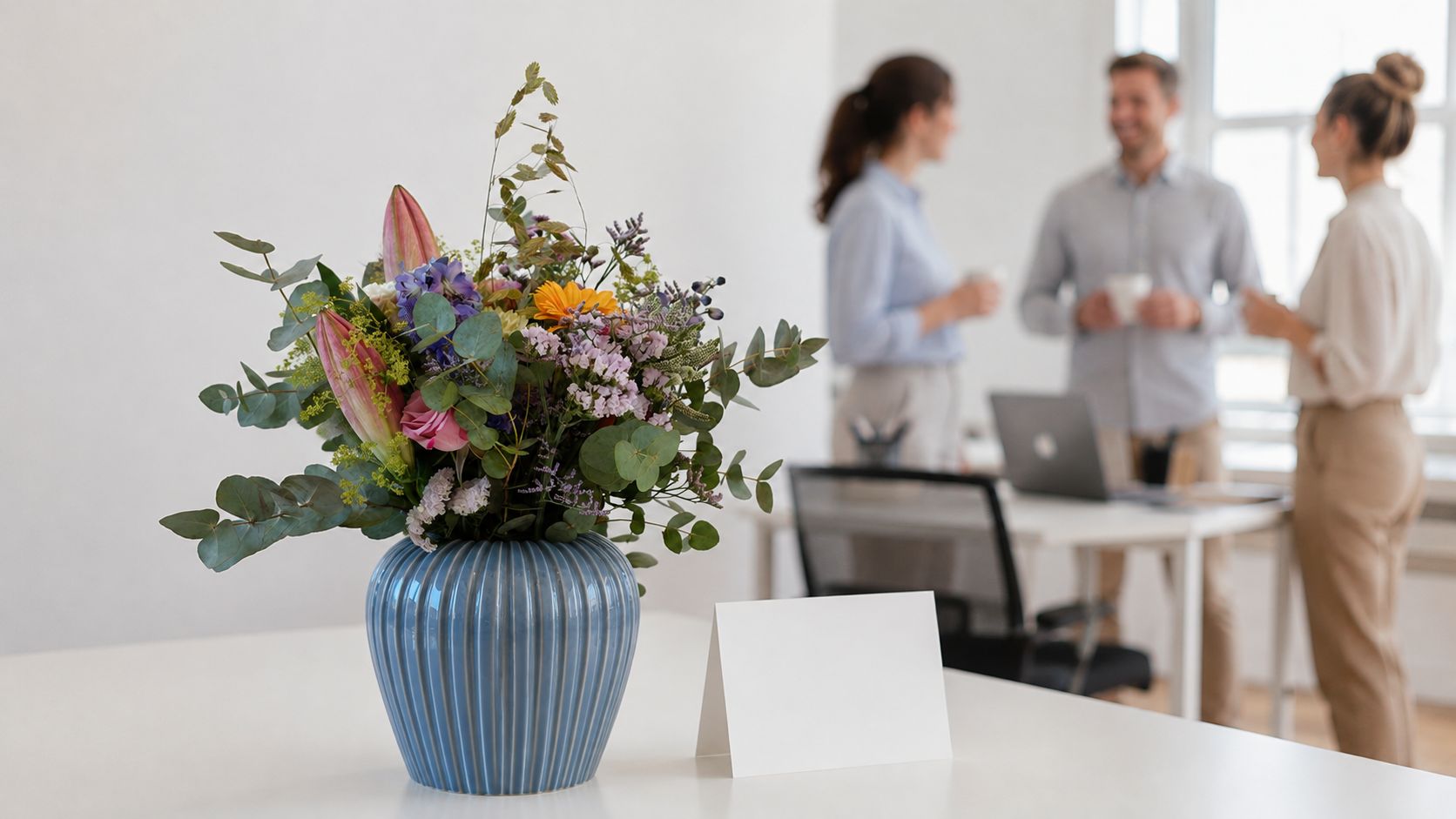 En smuk buket blomster i en blå vase står på et bord ved siden af et tomt lykønskningskort.