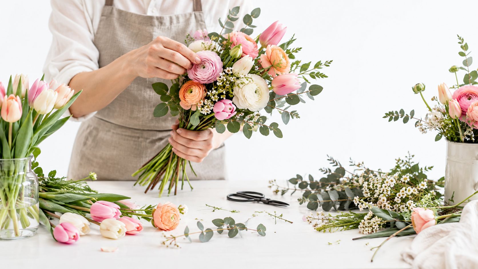 En florist arrangerer en smuk buket med forårsblomster, tulipaner og ranunkler ved et arbejdsbord i et lyst værksted.