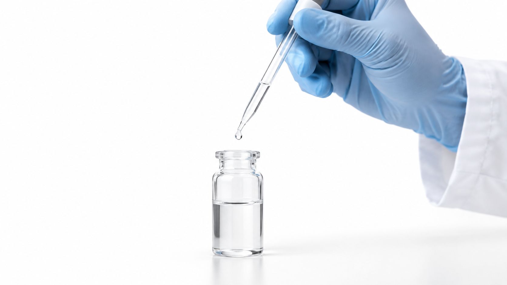 A gloved hand uses a glass dropper to add liquid into a small clear medicine vial.