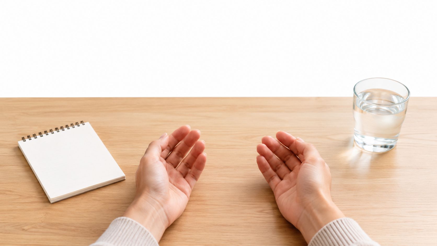 A person sitting at a wooden desk with a notebook and glass of water, showing open palms.