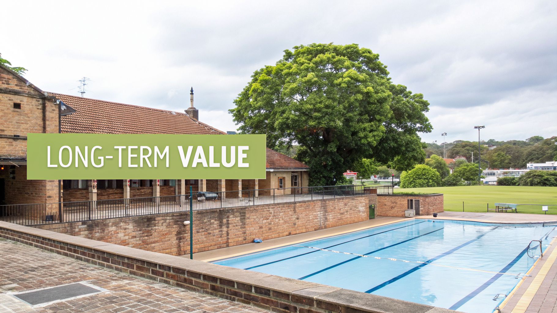 An outdoor swimming pool with blue lanes, a brick building, and a large green tree under a cloudy sky.
