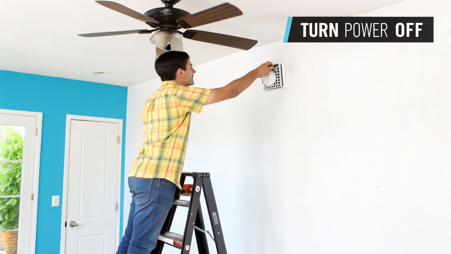 A man on a ladder reaching up to turn off power at a wall control panel near a ceiling fan.
