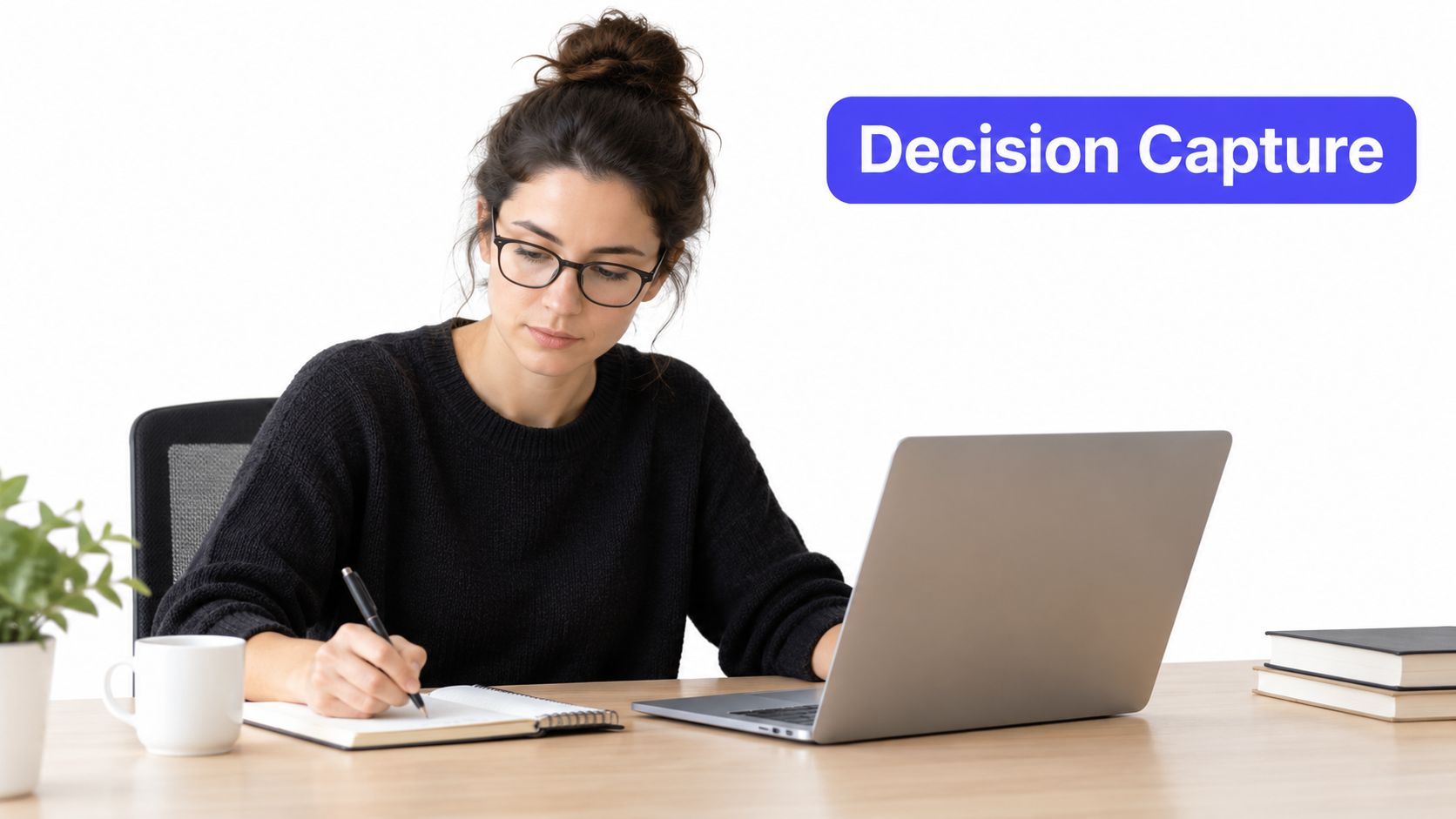 A woman wearing glasses working at her desk with a laptop and writing in a notebook.