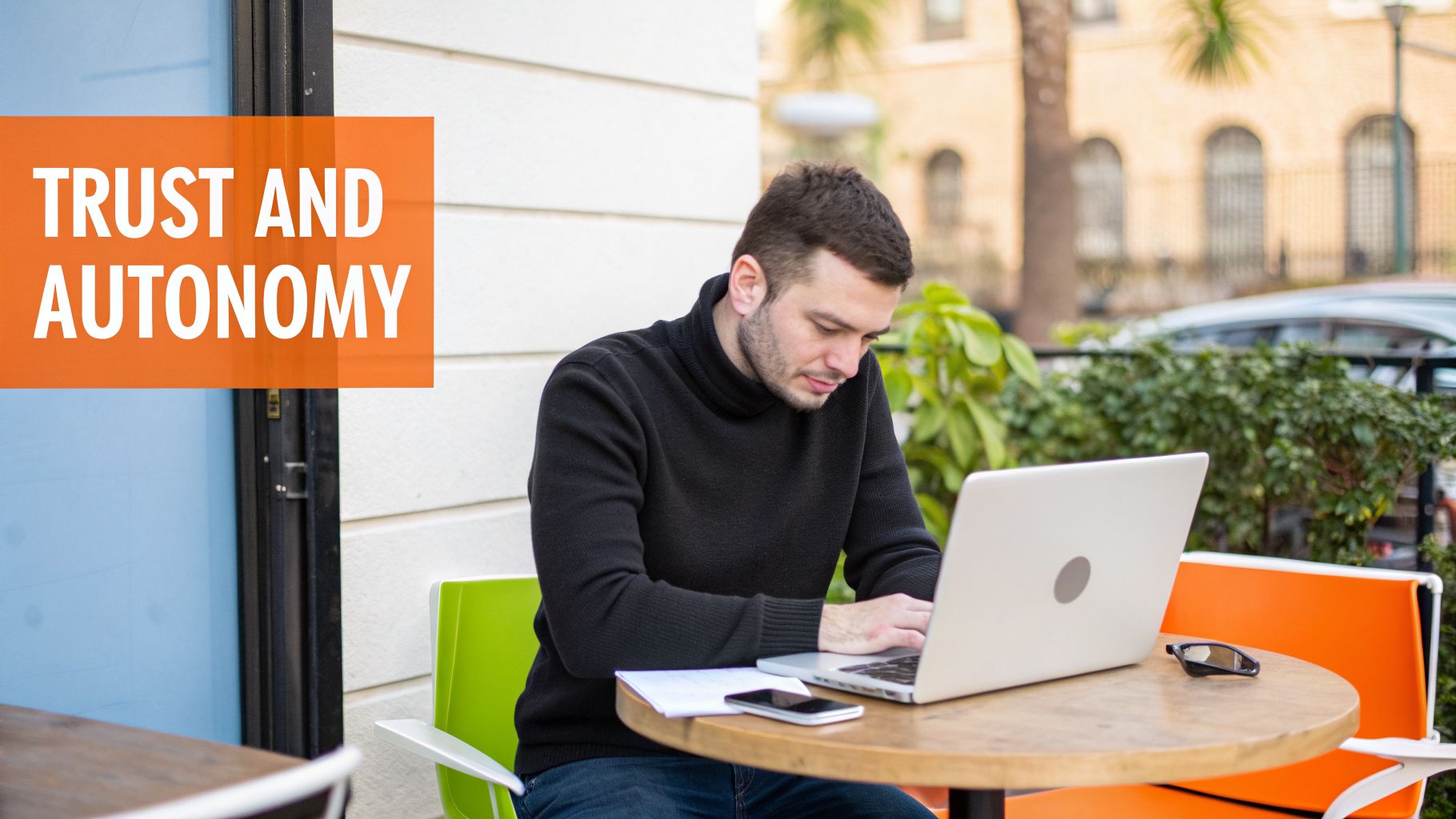 A man in a black turtleneck works on a laptop at an outdoor cafe with text 'TRUST AND AUTONOMY'.