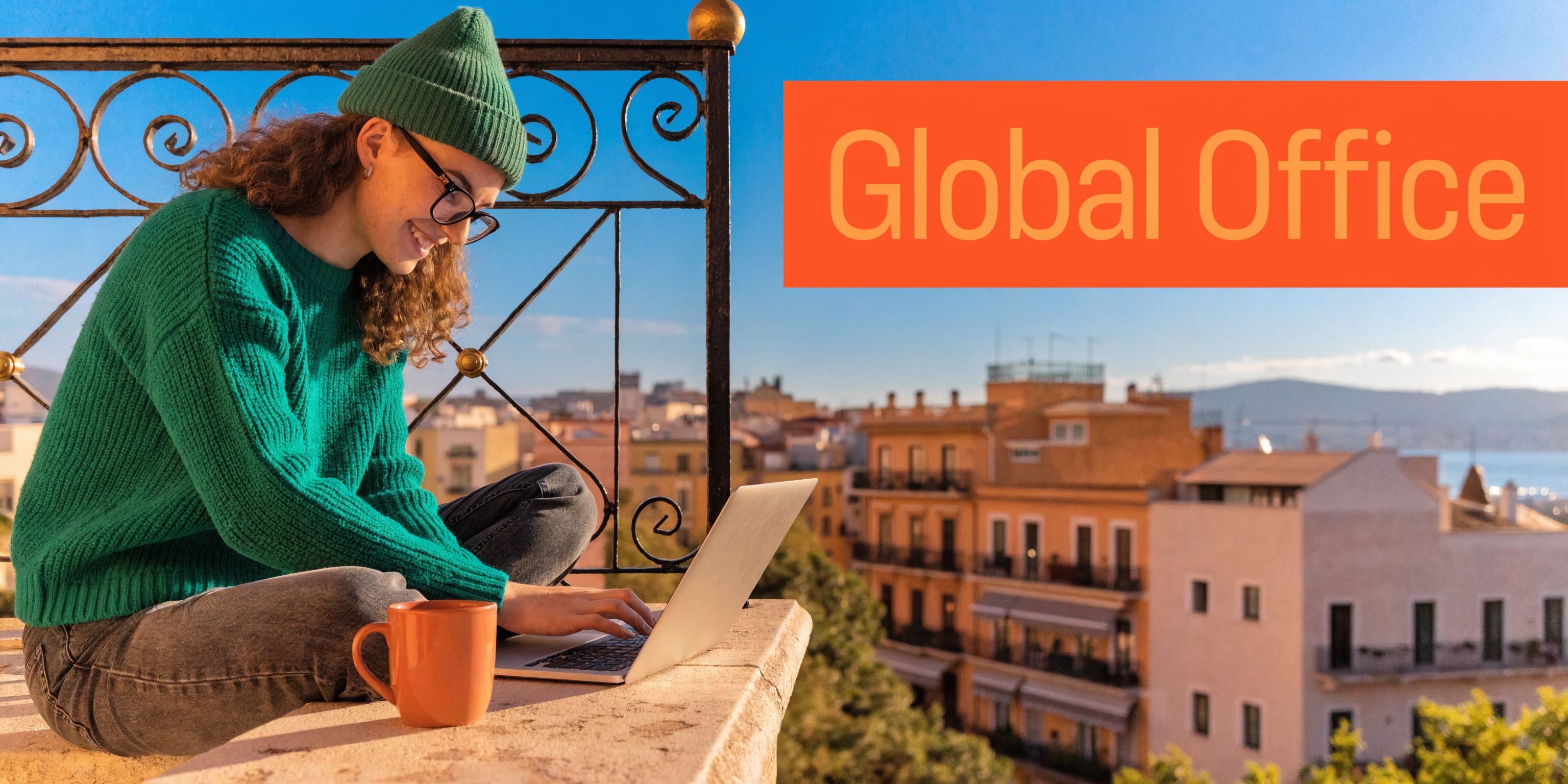 A happy person wearing a beanie working on a laptop on a sunny rooftop overlooking a city.