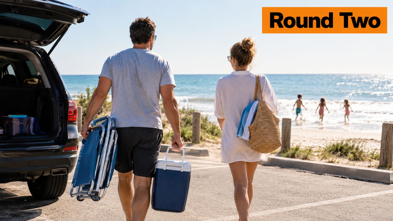 A couple walks towards the beach carrying a foldable chair, a cooler, and a straw bag.
