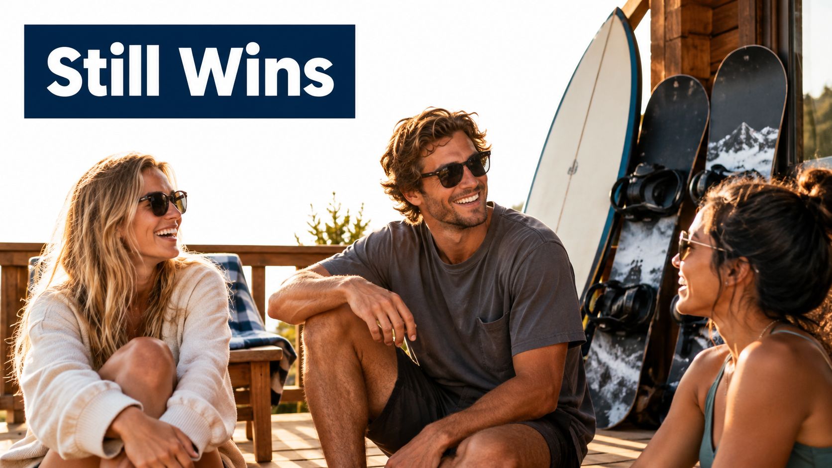 A smiling man and two women wearing sunglasses sitting on a wooden deck with surfboards and snowboards.