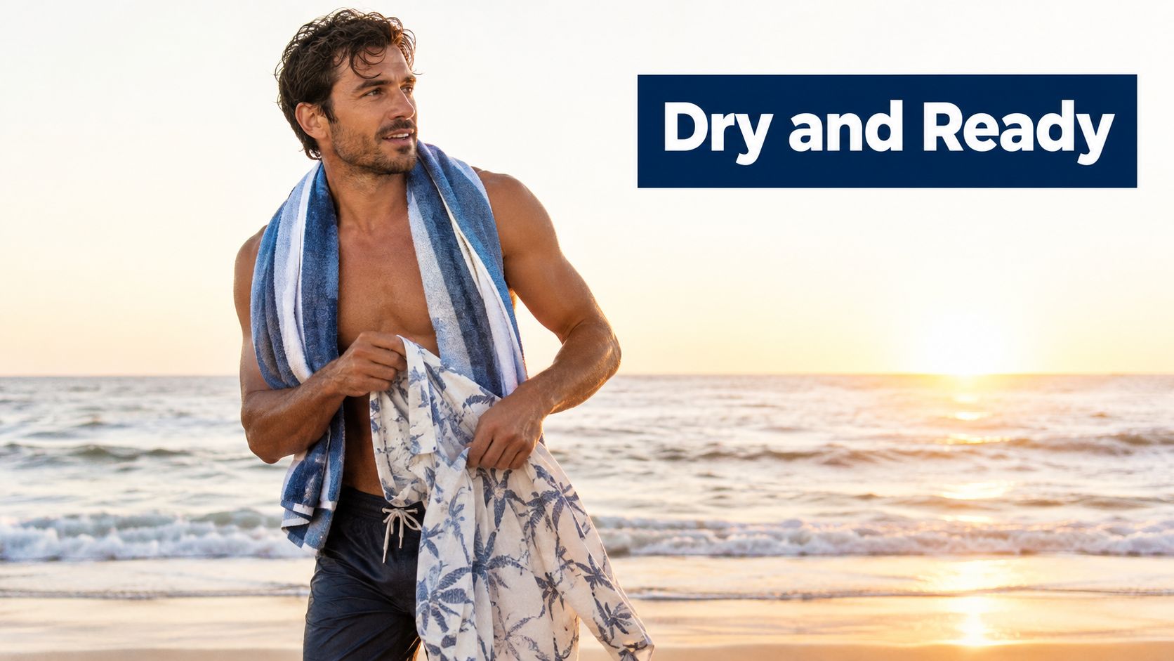 A fit man wearing swimming trunks holding a beach towel while standing on the sand at sunset.