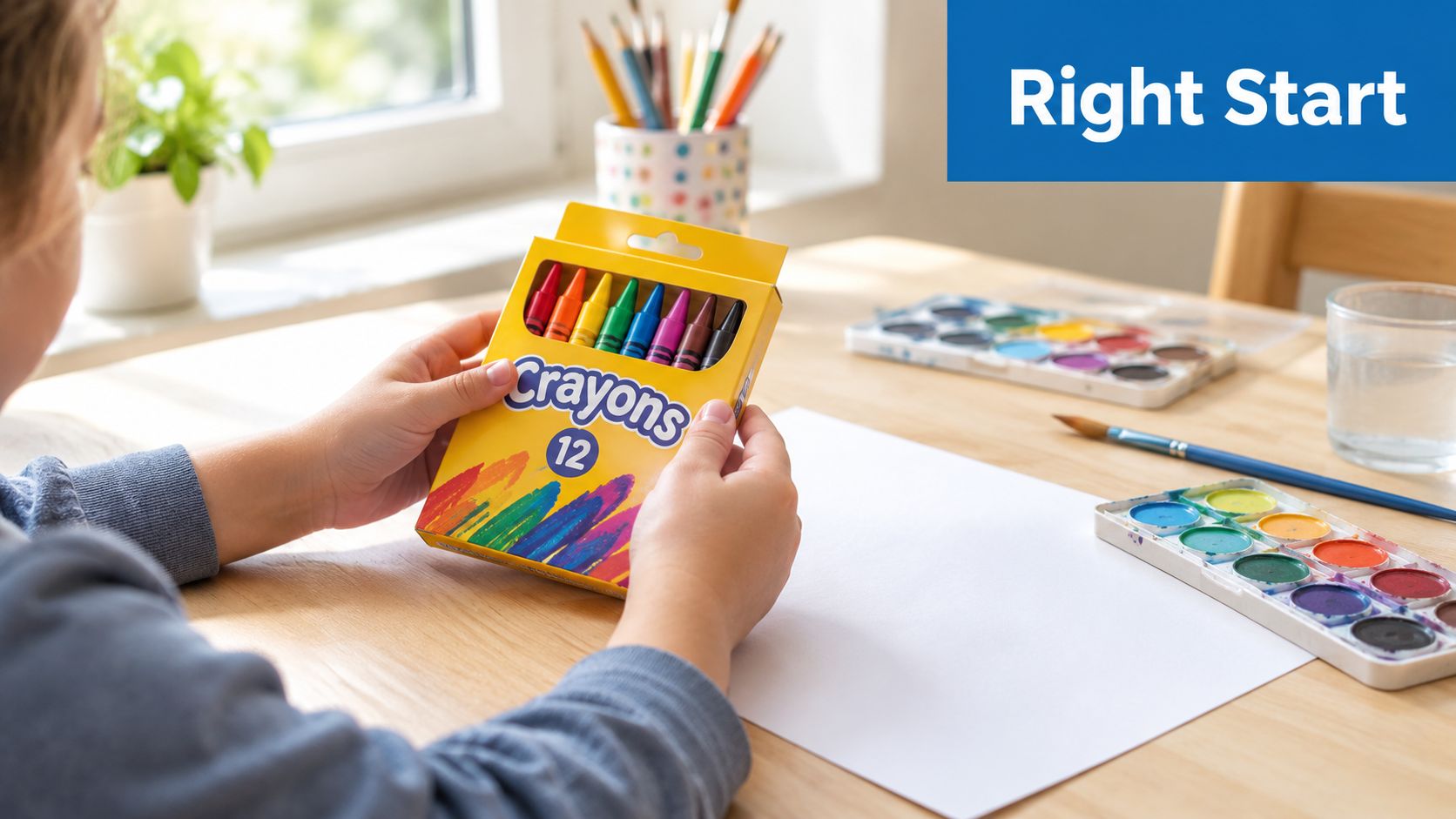 A young child holds a box of twelve colorful crayons while sitting at a wooden art desk.