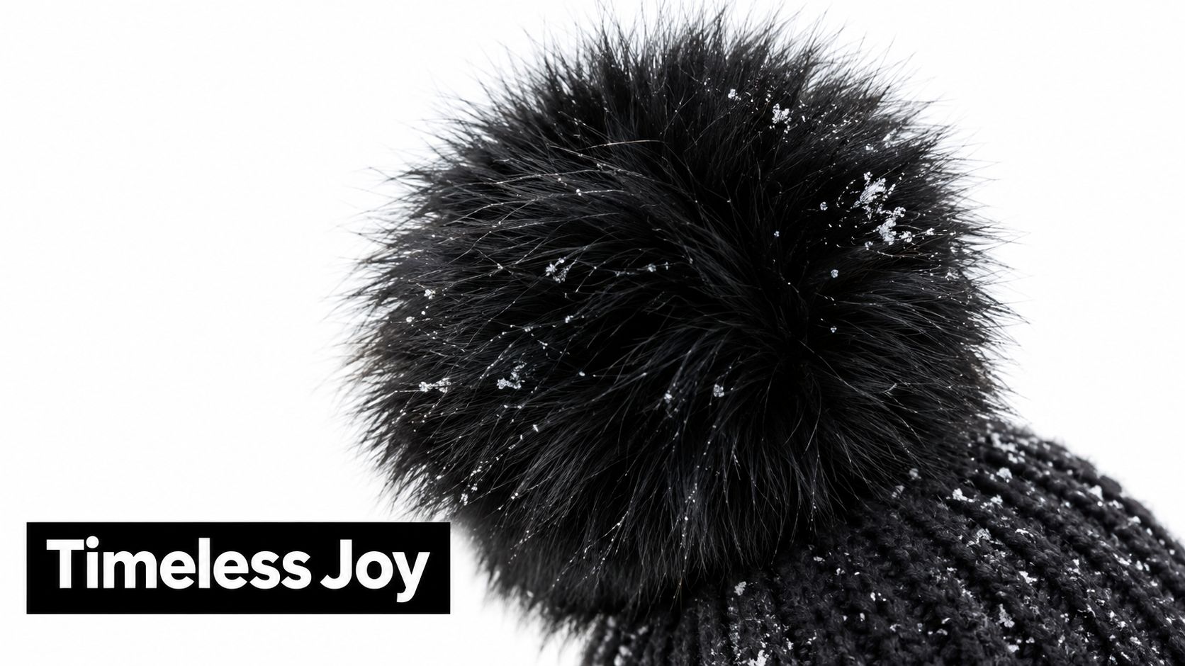 A close-up view of a black knitted winter hat with a fuzzy faux fur pom pom covered in snowflakes.