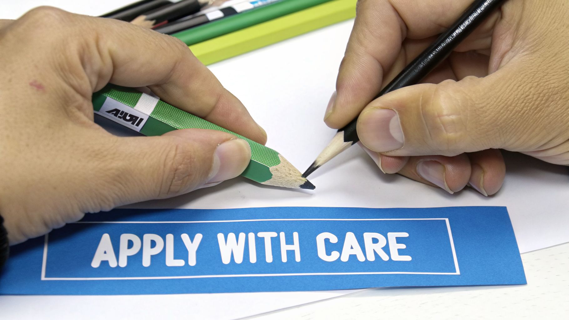 Two hands hold sharpened pencils with their tips nearly touching, above a blue label that reads 'APPLY WITH CARE'.