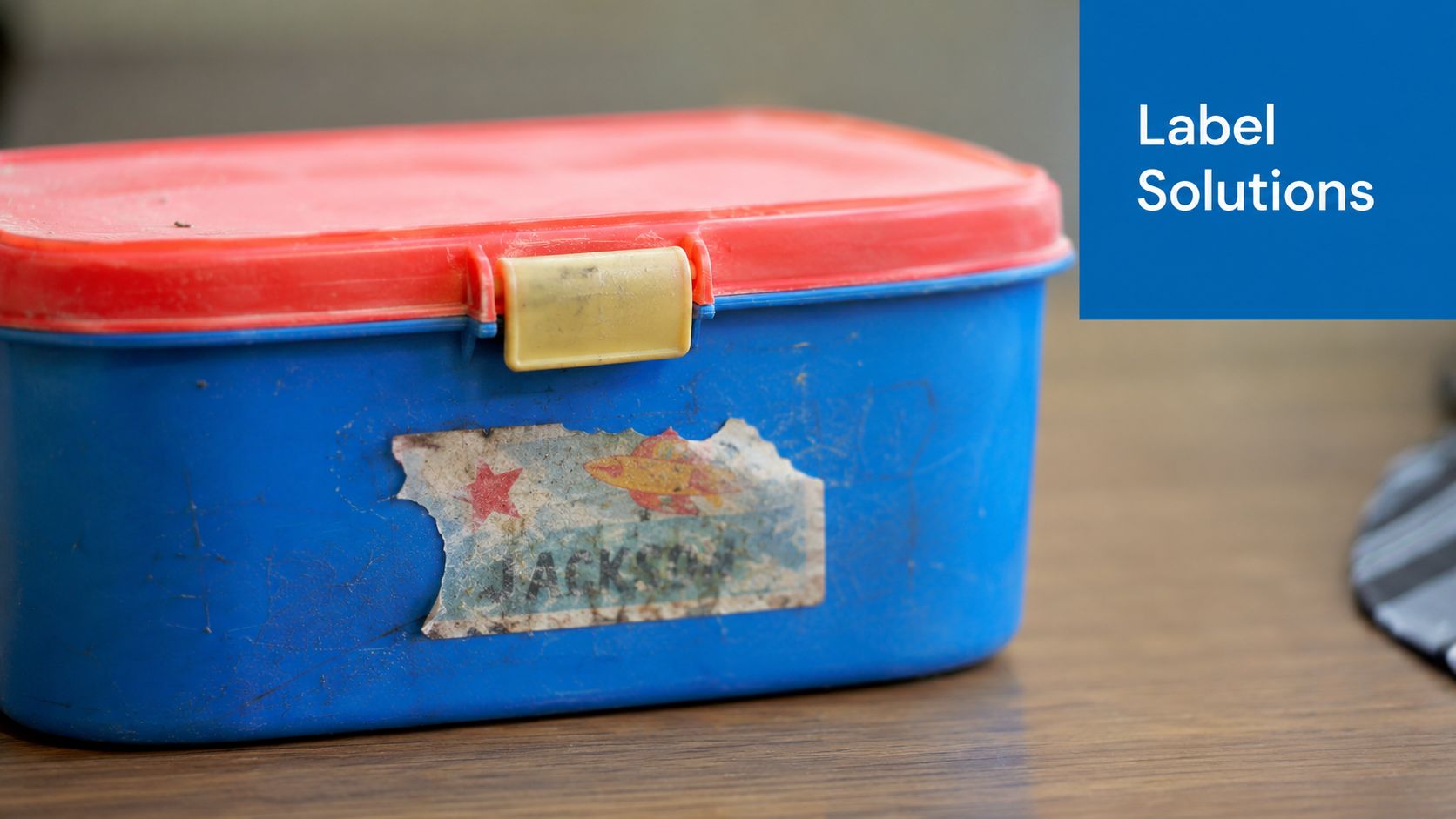 A worn blue lunchbox with a faded spaceship sticker and the name Jackson on a wooden table.