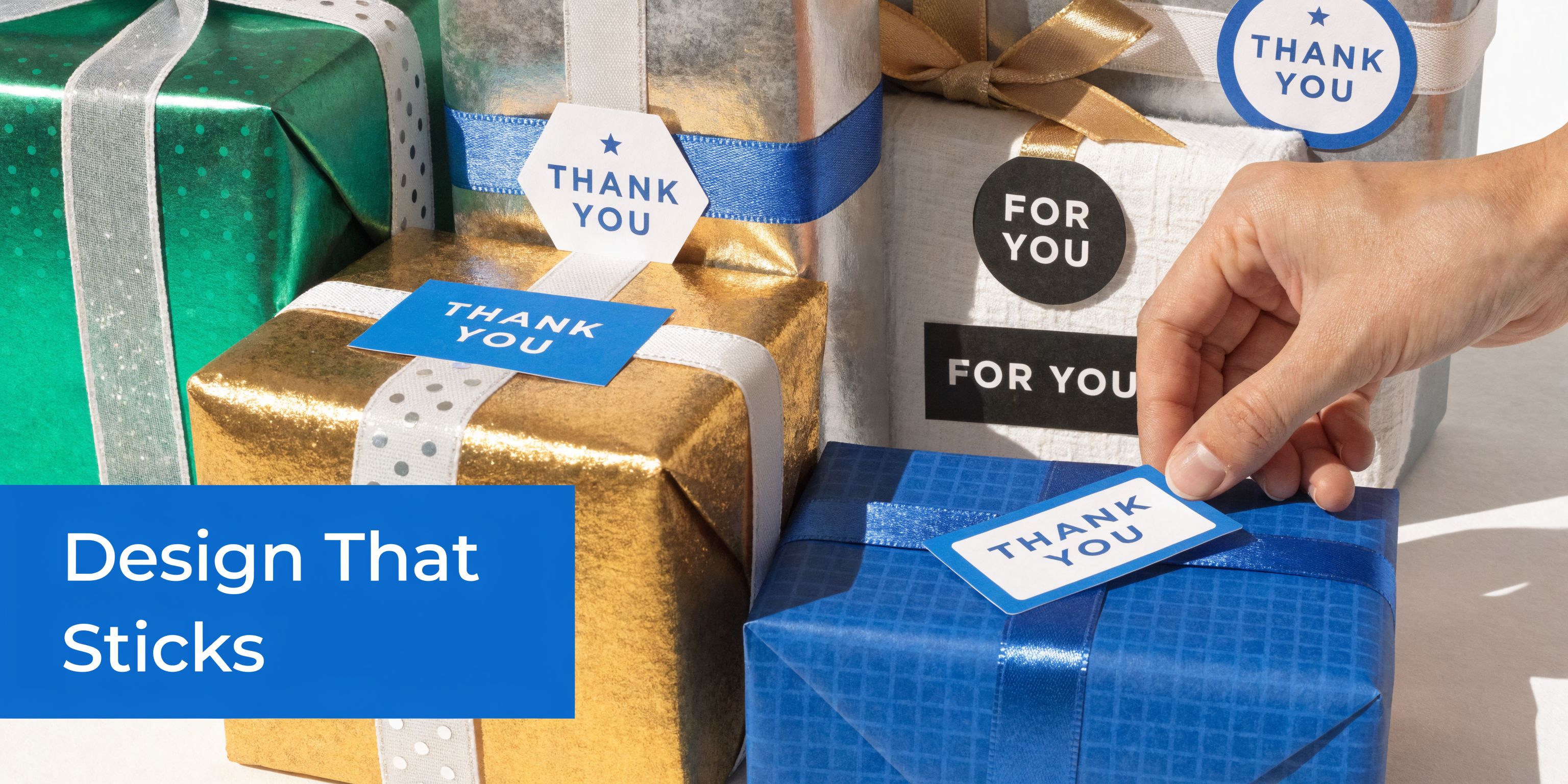 A hand placing a thank you gift tag sticker on a blue wrapped present with other gifts.