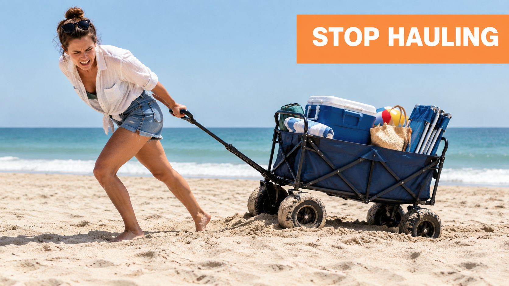 A woman struggling to pull a loaded beach wagon across loose sand on a sunny beach.