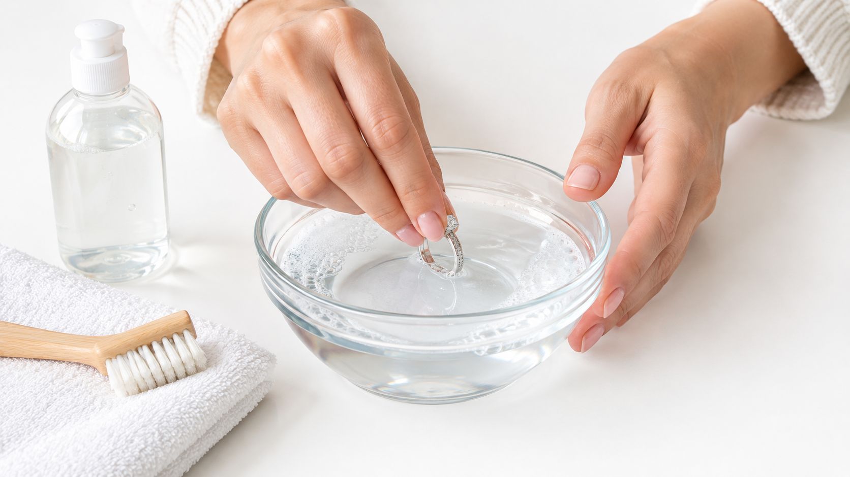 A person carefully cleans a diamond engagement ring in a bowl of soapy water at home.