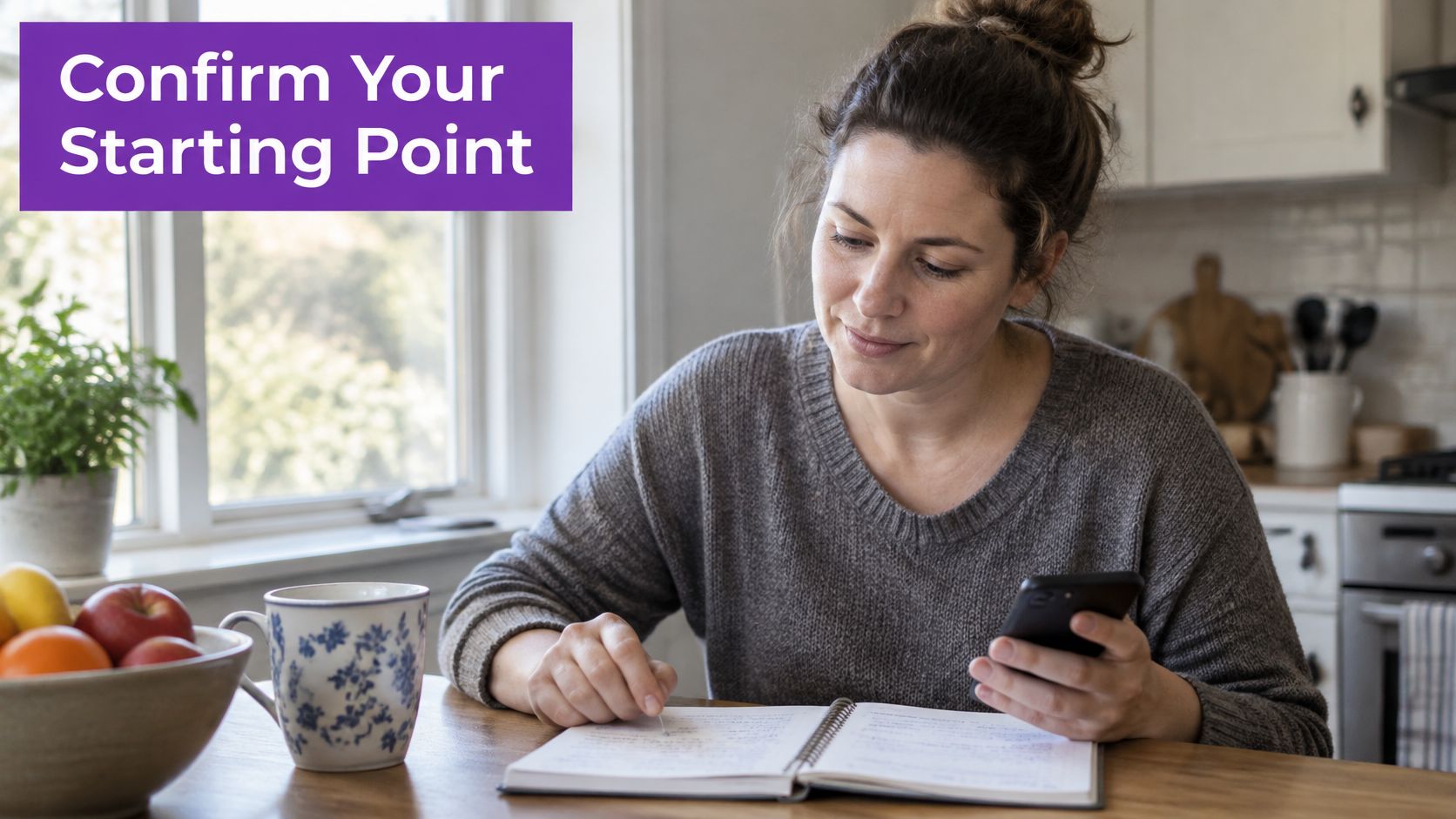 A woman sits at a kitchen table, looking at her smartphone while writing in an open notebook.