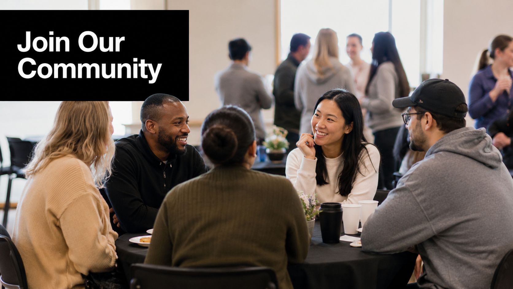 A diverse group of young adults sit around a table engaged in a friendly, smiling conversation.