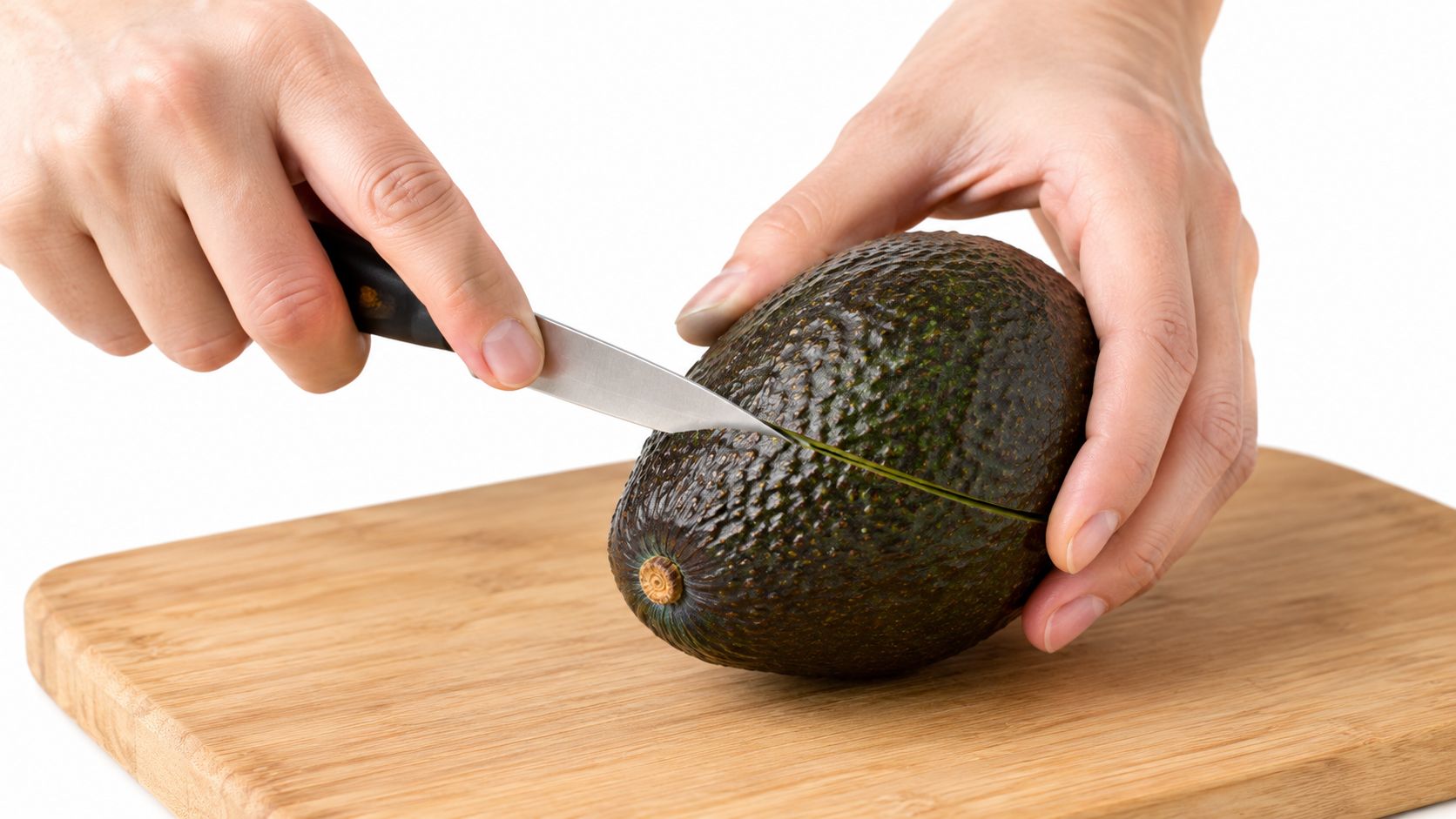 A pair of hands using a sharp knife to slice a fresh, ripe avocado on wood.