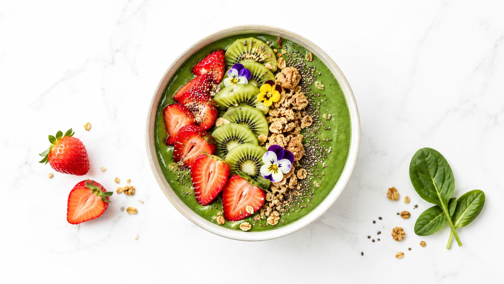 A vibrant green smoothie bowl topped with fresh sliced strawberries, kiwi, granola, chia seeds, and edible flowers.