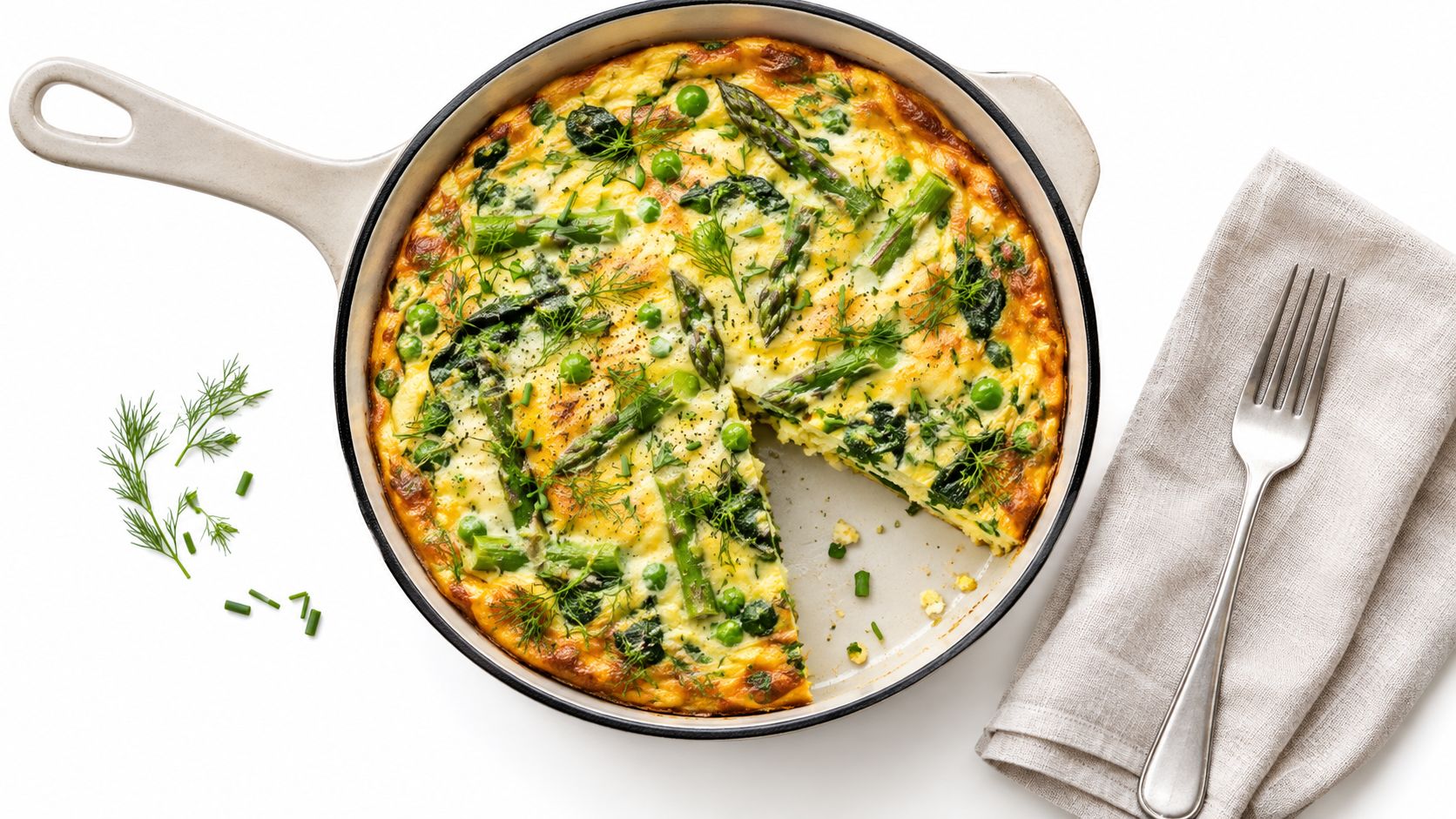 A freshly baked vegetable frittata with asparagus, peas, and dill in a white skillet on a table.