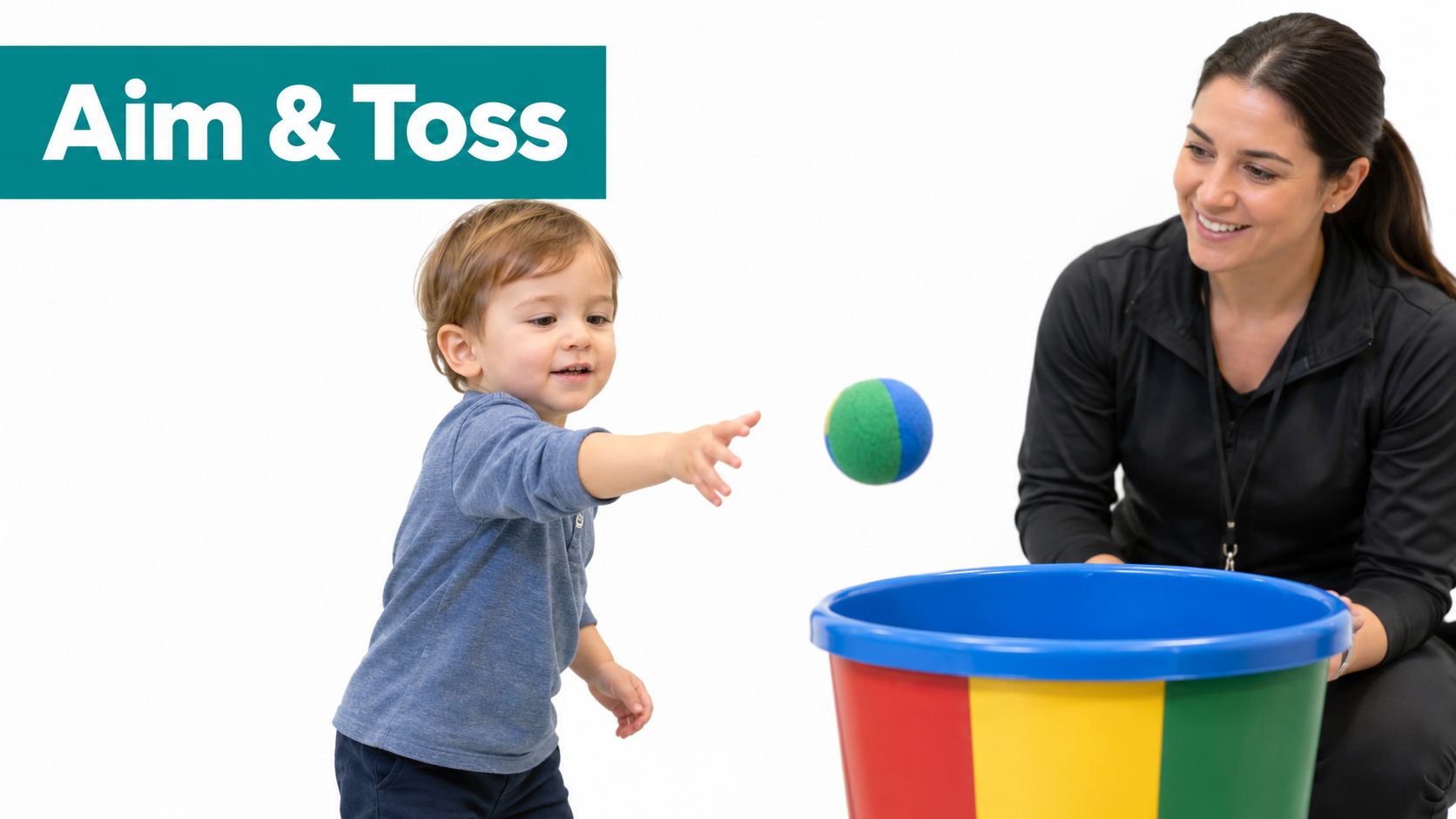 A smiling toddler throws a soft ball into a colorful bucket while a woman watches nearby.